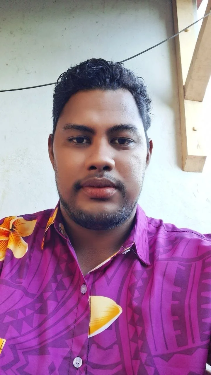 A man with dark curly hair and a beard taking a selfie, wearing a purple shirt with yellow and orange floral patterns. He is sitting outdoors near a wall with a wooden beam and a visible wire.