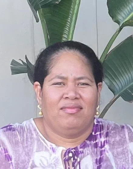 A woman with dark hair pulled back, wearing earrings and a purple and white patterned top, standing outside in front of large green leaves.