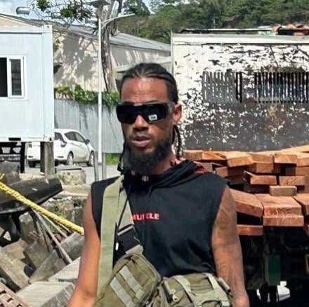A man with long hair and a beard wearing sunglasses, a black sleeveless hoodie, and carrying a bag, standing outdoors near construction materials and a parked car.