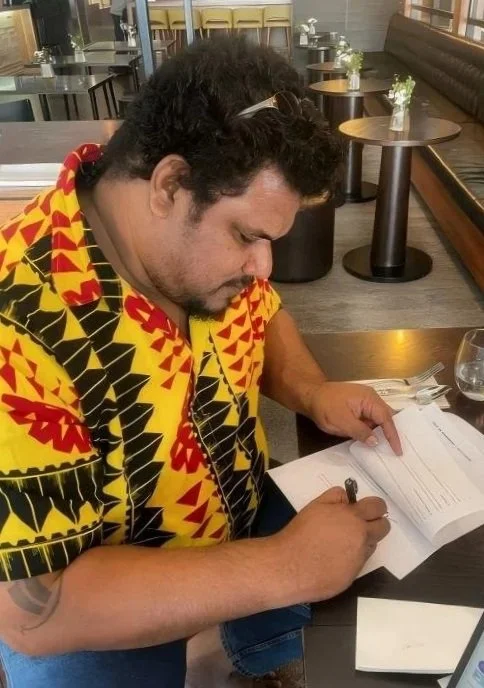 A man with curly hair and a mustache wearing a colorful, patterned shirt, sitting at a table in a restaurant, writing in a notebook.