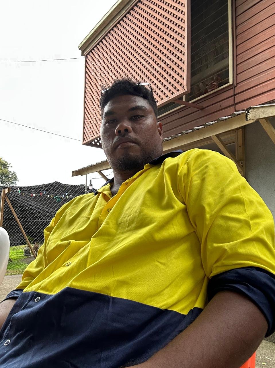 A man in a yellow and navy blue shirt sitting outdoors, with a wooden house and a fenced yard in the background.