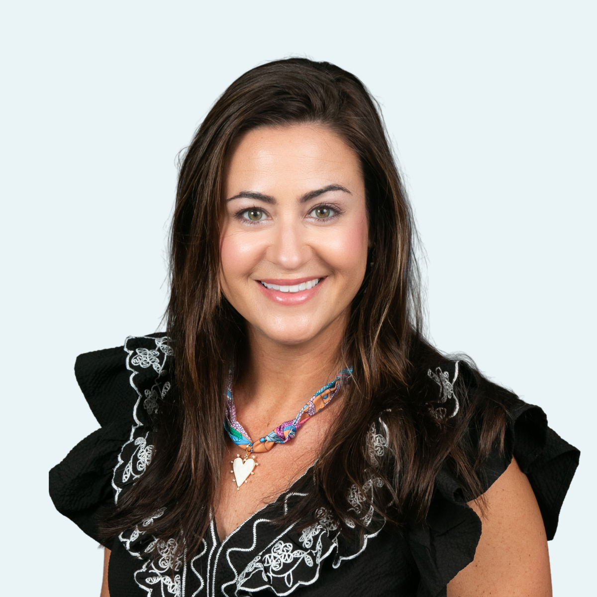 A smiling woman with brown hair and green eyes wearing a black top with ruffled sleeves, a colorful necklace with a heart pendant, and a light blue background.