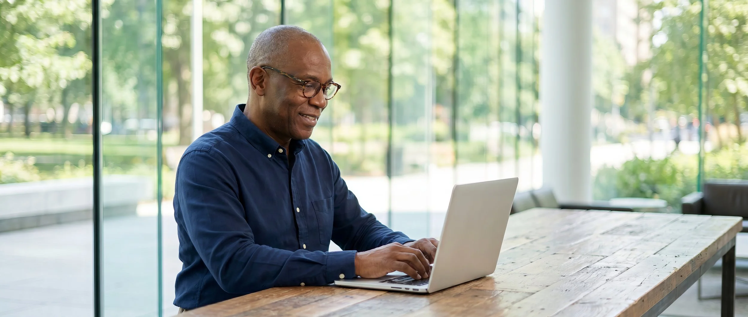 An older man with glasses sitting at a wooden table, using a laptop in a bright room with large windows and a view of green trees outside.