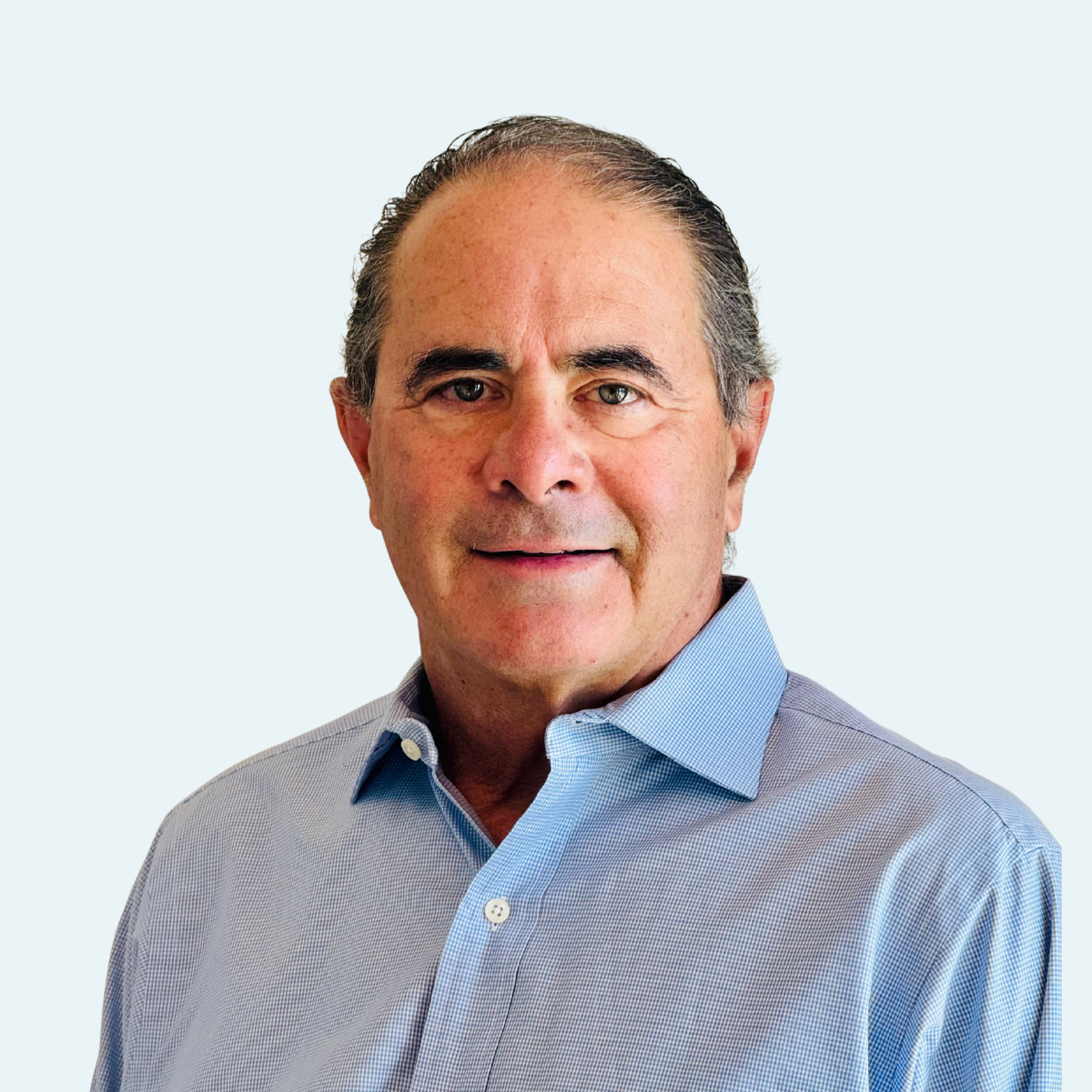 Close-up portrait of a middle-aged man with gray hair, wearing a blue button-up shirt, looking at the camera against a light background.