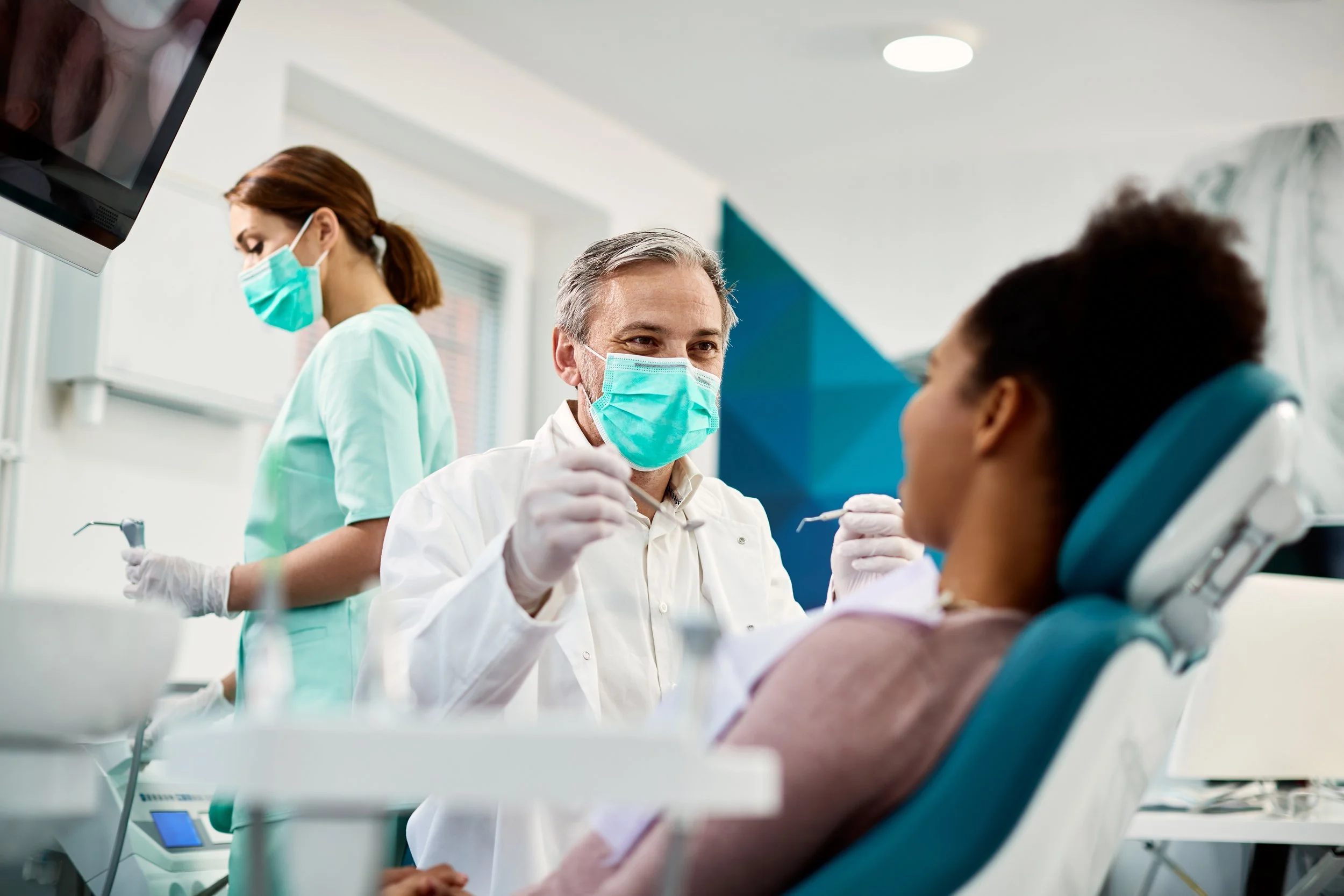 A young girl lying in a dental chair, a male dentist wearing a face mask and gloves examining her teeth, an assistant in the background also in a mask, monitoring a screen in a dental office.
