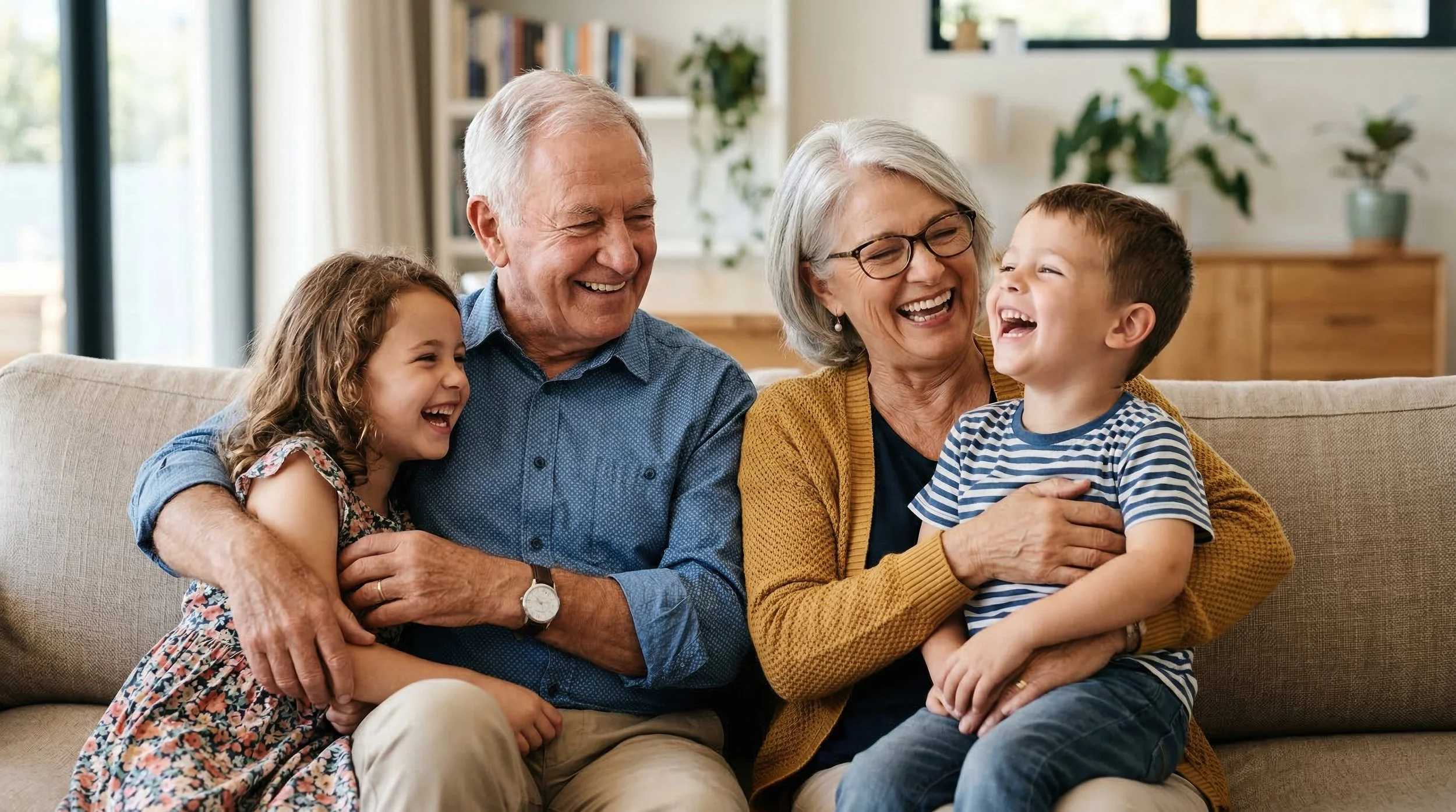An elderly man and woman, with two young children, sitting on a couch and laughing together in a bright living room.