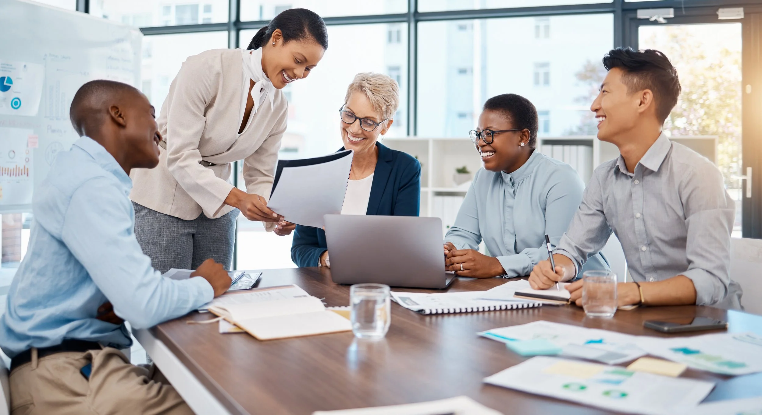 A diverse group of six business professionals in a meeting room, sitting around a table with documents, a laptop, and glasses of water, smiling and engaging in discussion.