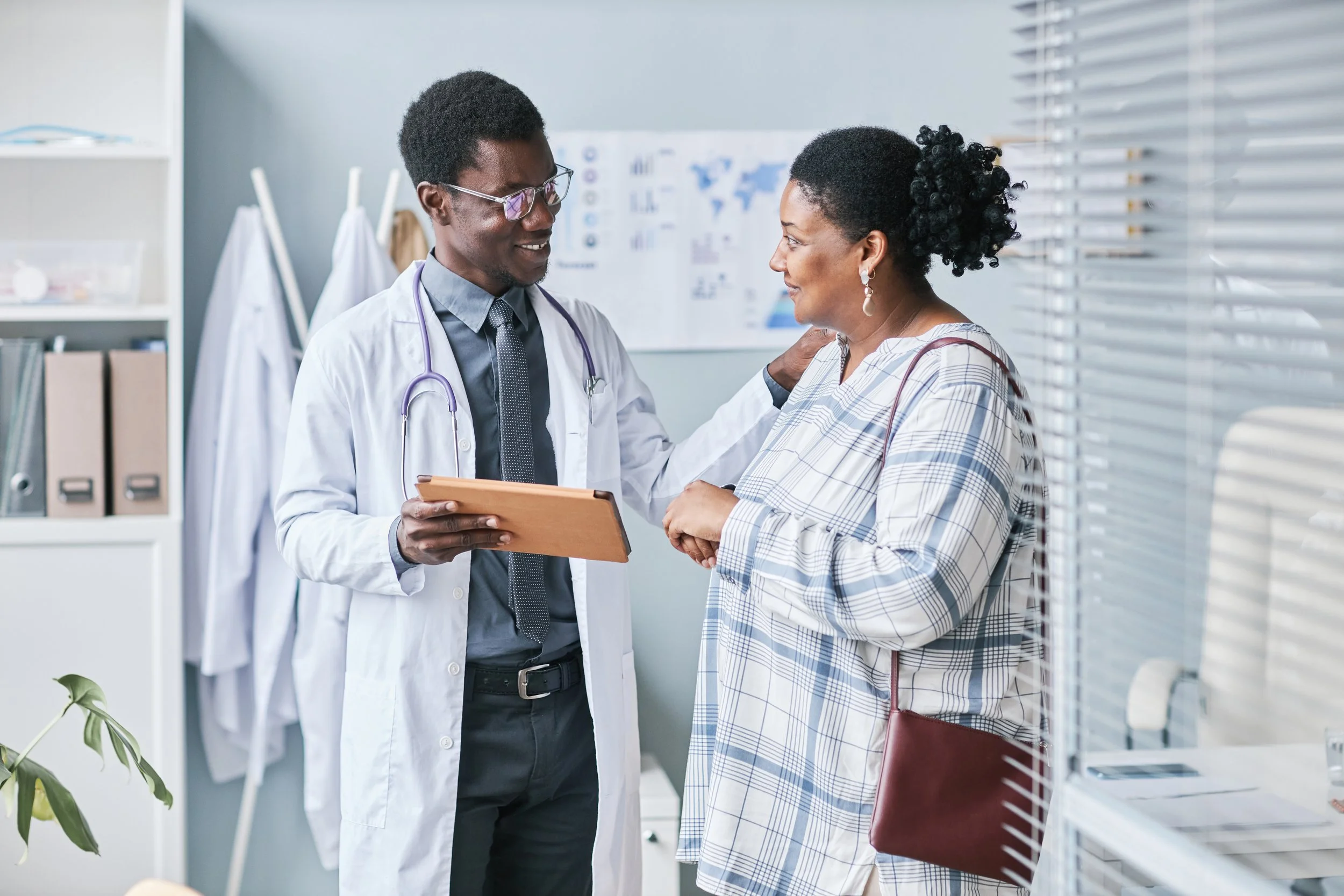 A doctor and a woman share a moment in a medical office. The doctor, wearing a white coat, glasses, and a stethoscope, is smiling and holding a clipboard. The woman, wearing a plaid shirt, is smiling back. The background shows office supplies, medical charts, and a window with blinds.