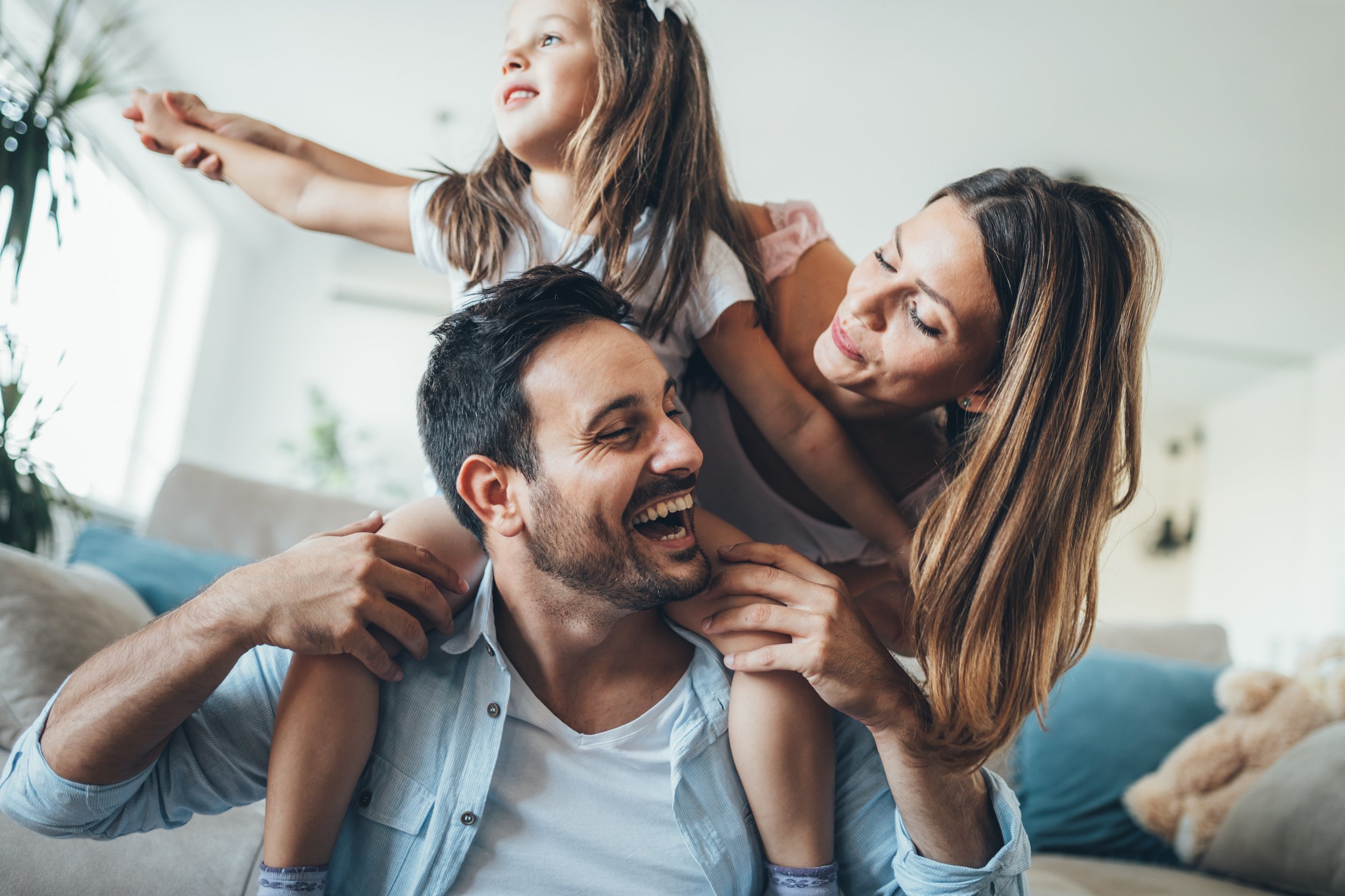 A family of three having fun together indoors. The man is sitting on a couch, smiling with eyes closed, holding the young girl on his shoulder, who is reaching out front. The woman is leaning in close to the man, smiling and touching his face.
