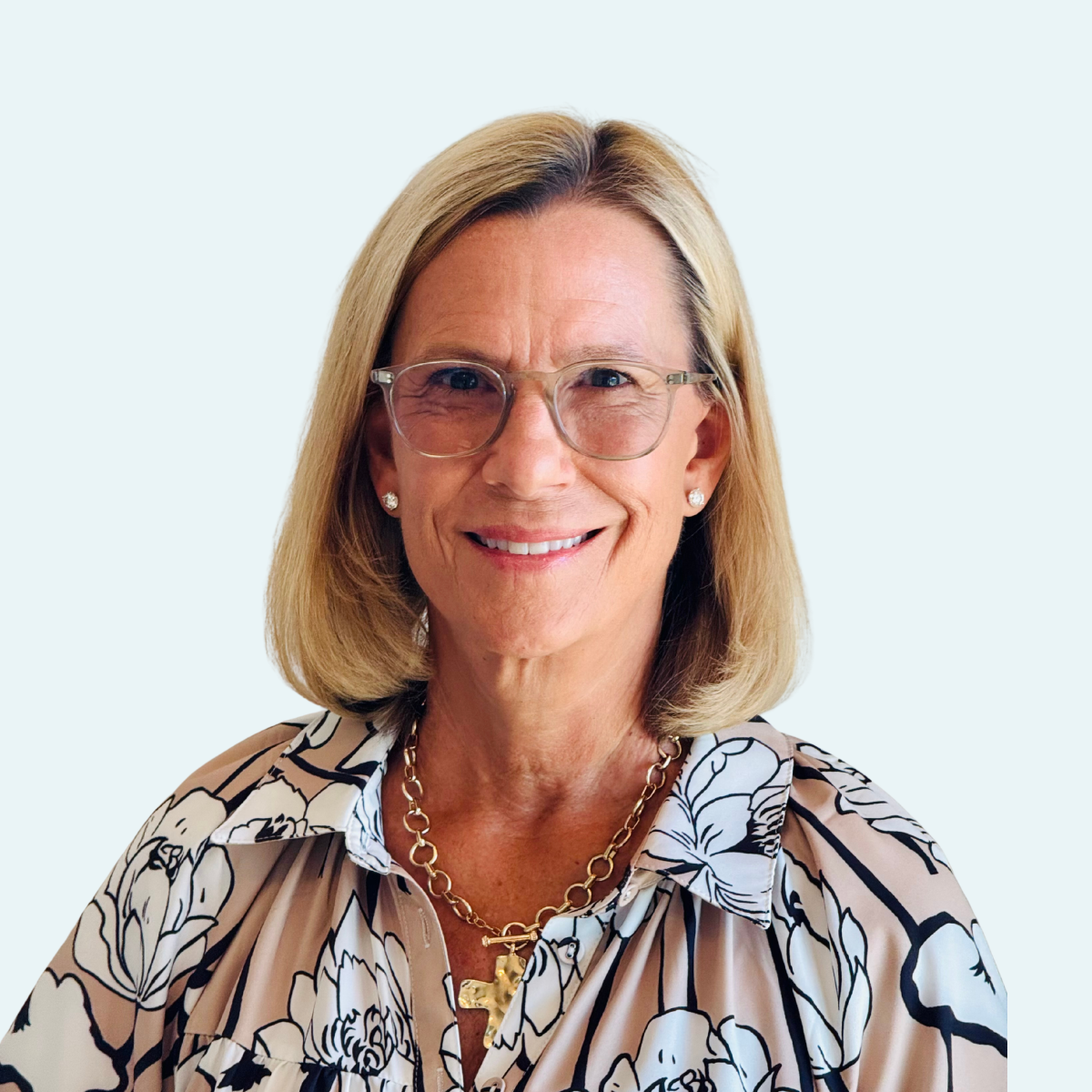 A smiling middle-aged woman with blonde hair and glasses, wearing a floral blouse, jewelry, and earrings, against a light blue background.