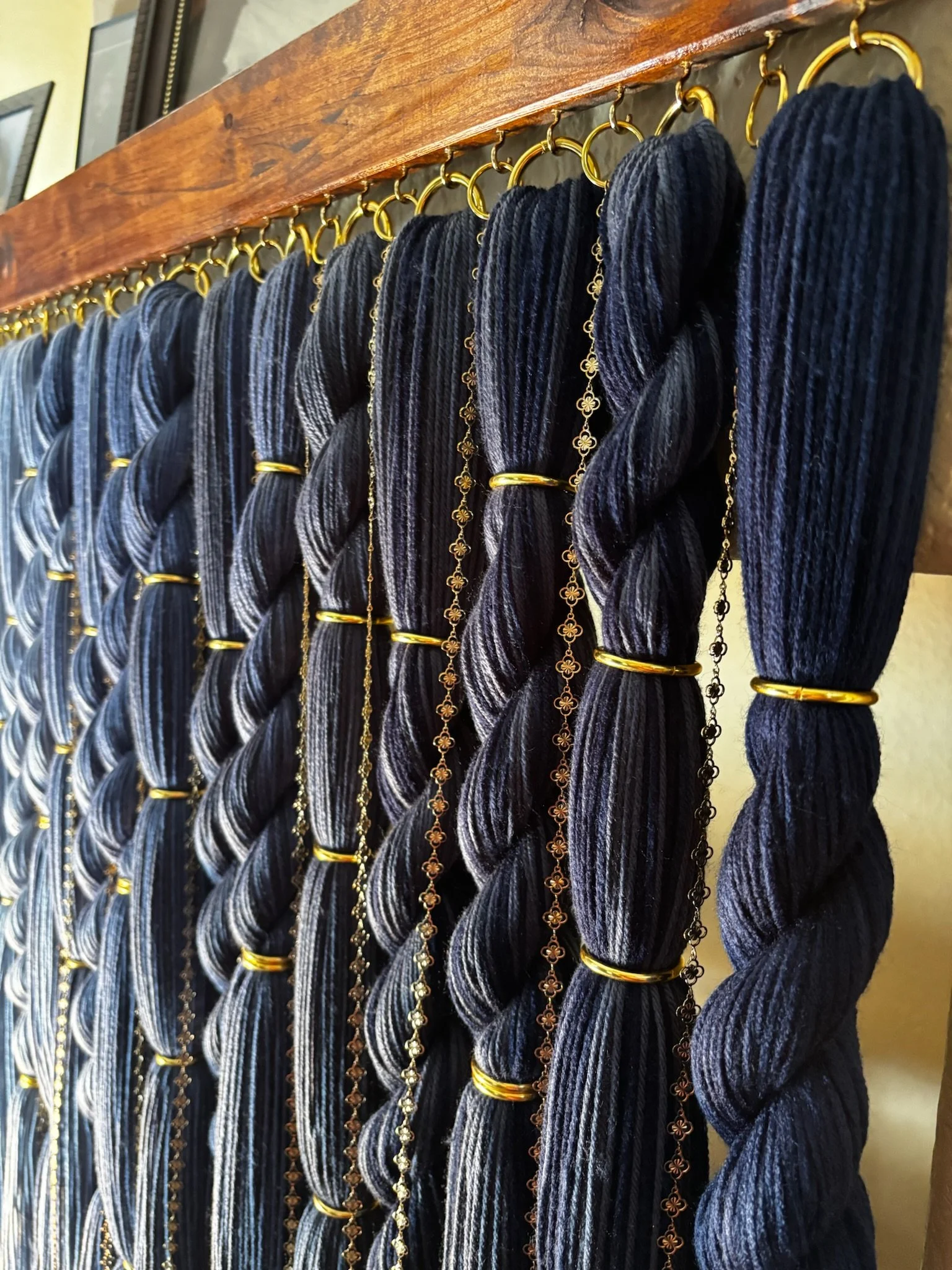 Display of various dark-colored yarn wall art hanging on a wood grain base secured with gold rings and chains, with a wooden shelf above.