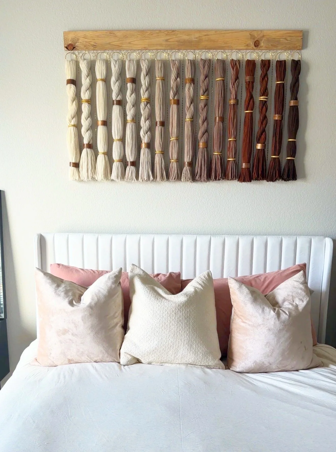 Decorative wall hanging with multicolored yarn strands above a white bed with multiple pillows.