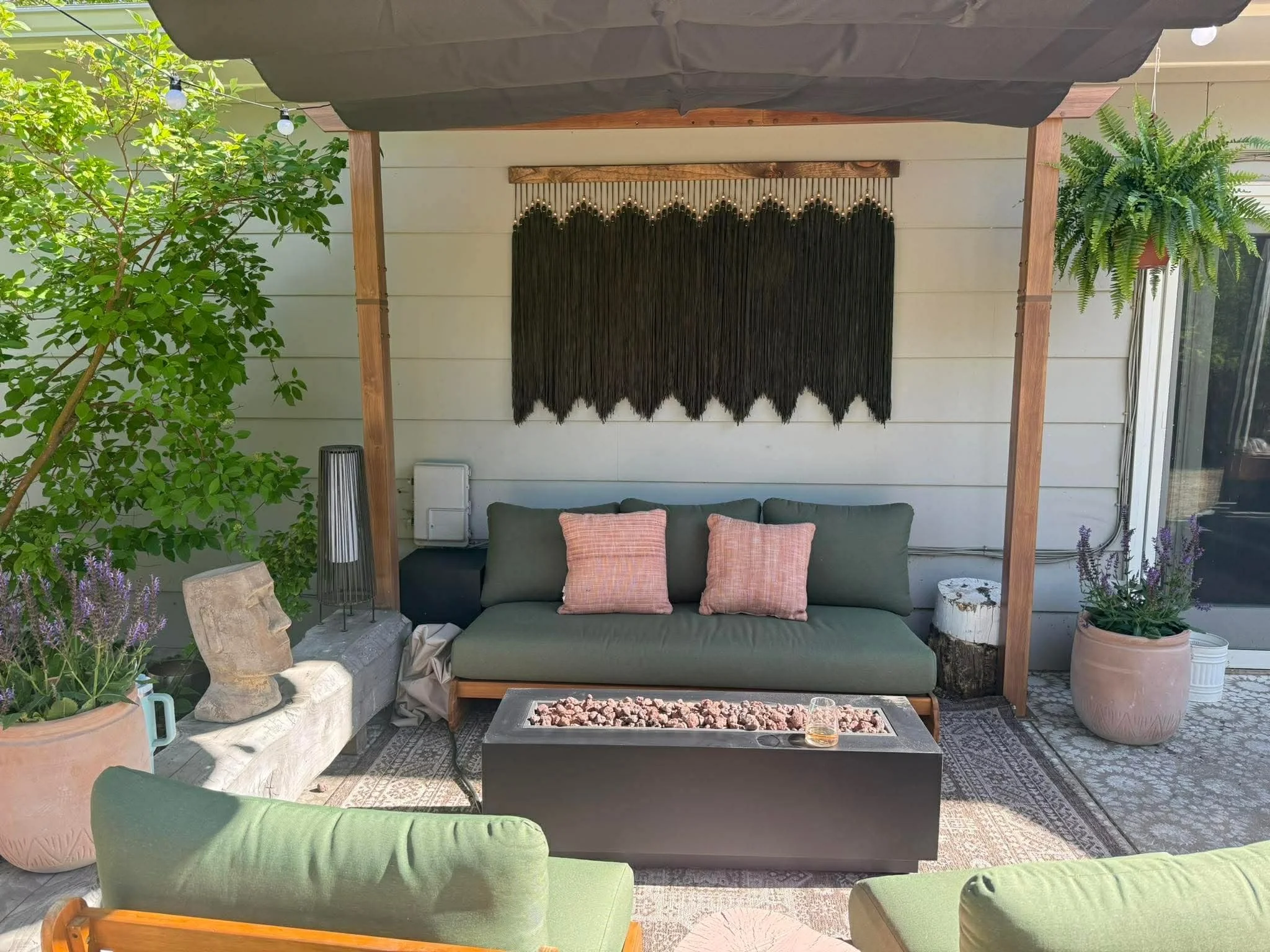 A cozy outdoor seating area with a green sofa, pink pillows, a small coffee table with rocks and a drink, surrounded by potted plants and a wooden pergola with black hanging macramé wall art.