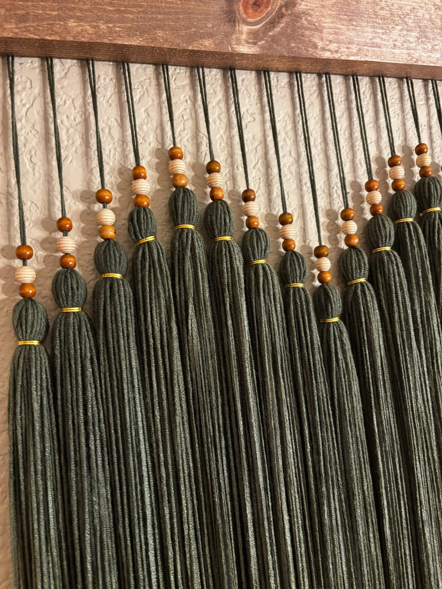 Decorative wall hanging with gray tassels, wooden beads, and small cream-colored beads hanging from black cords against a textured white wall and wooden top.