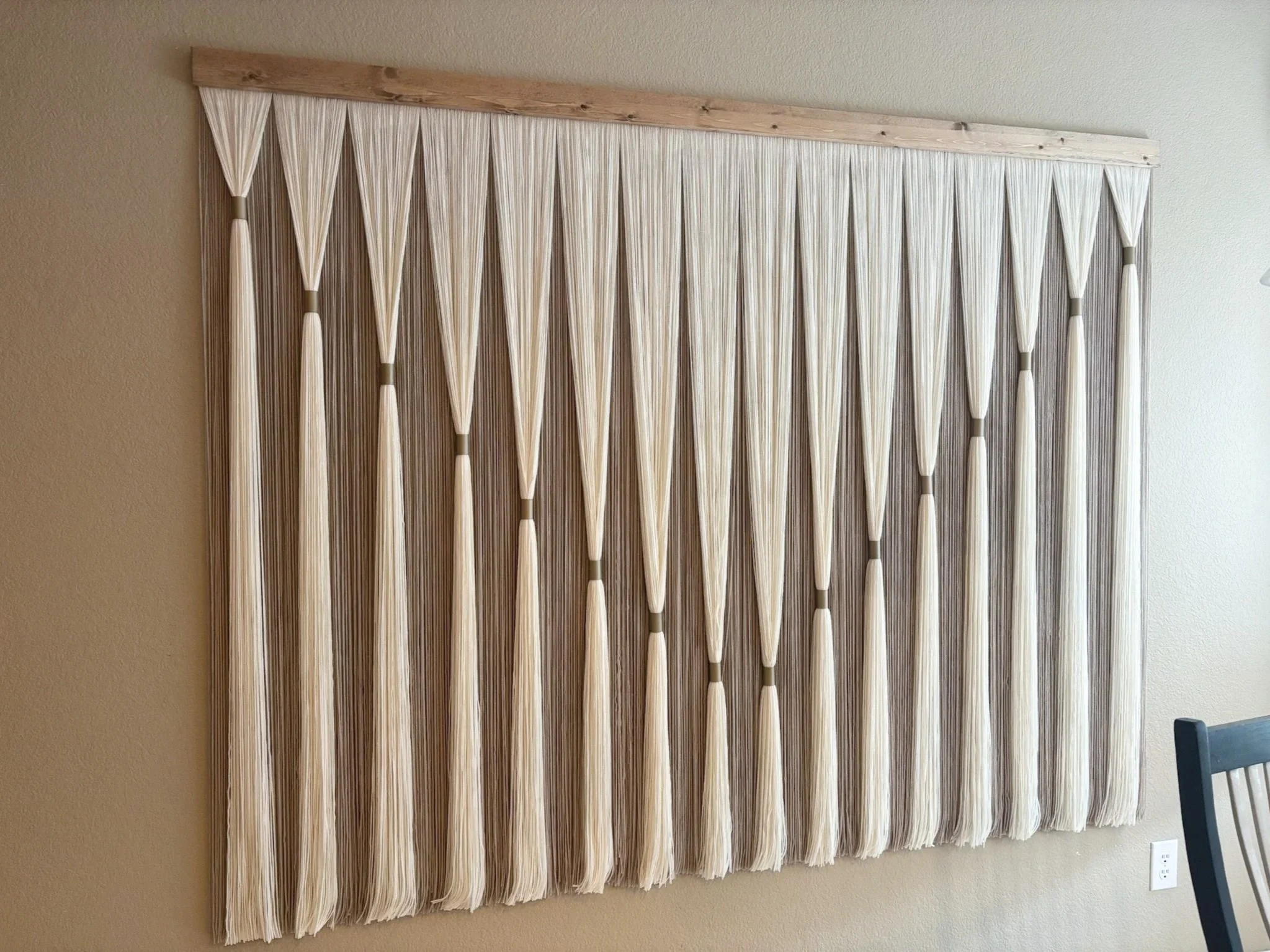 Wall hanging art made from white and beige strings arranged in a geometric pattern, with a wooden top rail, hanging on a beige wall next to a corner of a room.