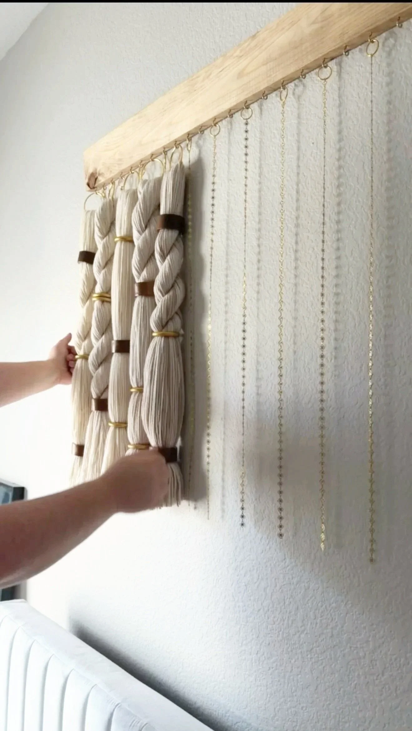 Hands installing or adjusting a decorative curtain or textile with twisted, pleated fabric on a wooden rod, on a textured white wall.