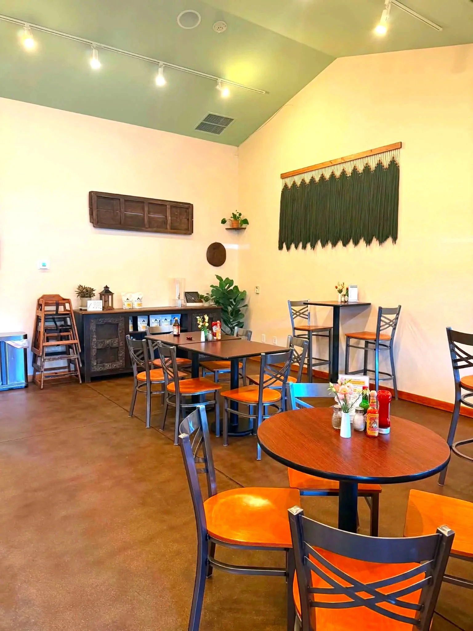 Inside a cozy cafe with wooden tables and chairs, green and yellow wall decorations, potted plants, and condiment bottles on the tables.