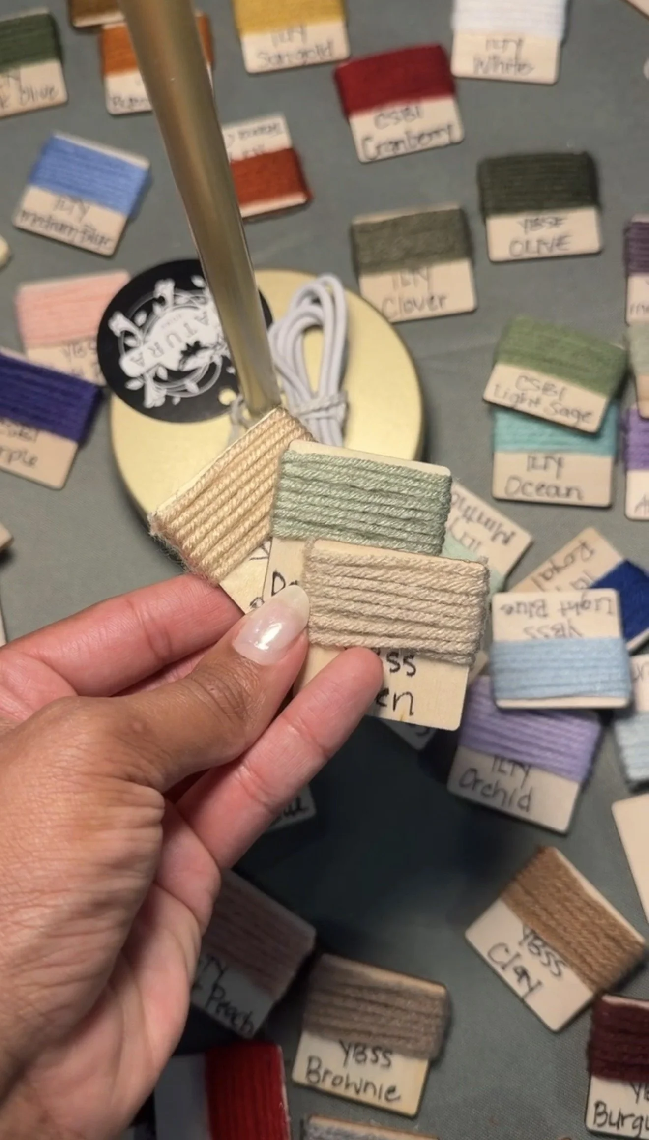 A hand holding three fabric color samples labeled "Yass" with names like "Brownie" and "Cream" amongst other fabric swatches on a table.