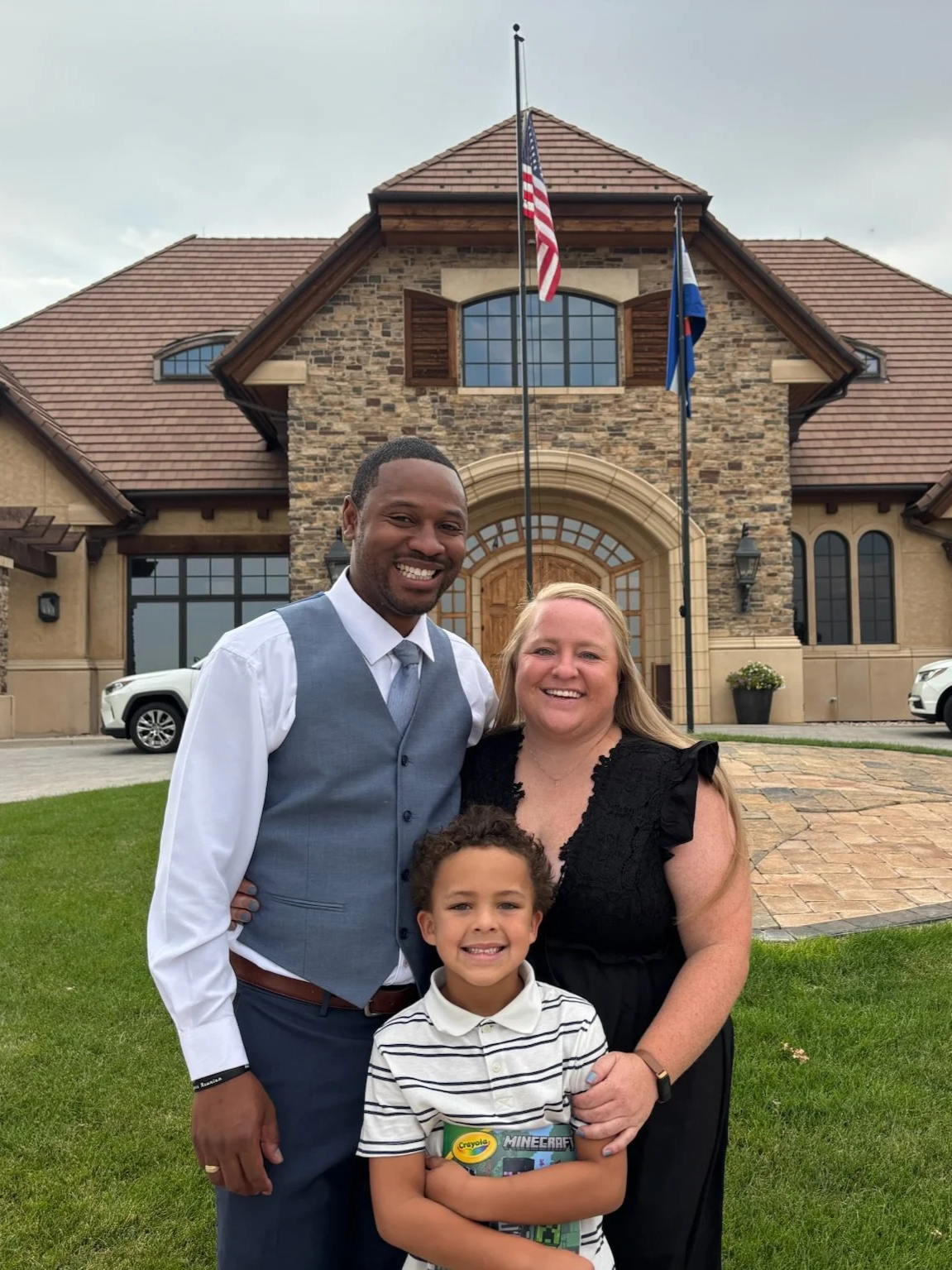 A family of four smiling in front of a large house with American and state flags. The man is wearing a blue vest and white shirt, the woman has long blonde hair and is wearing a black dress, the boy is holding Crayola and Minecraft boxes, and the younger boy is smiling in front of them.