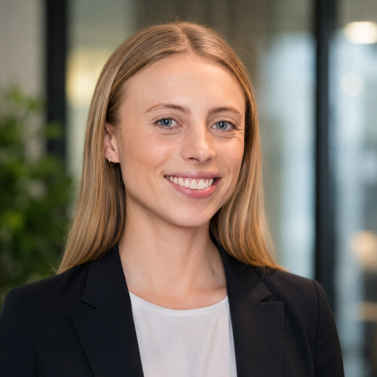 A young woman with long blonde hair, blue eyes, and a bright smile, wearing a black blazer and white top, standing indoors with a blurred background.