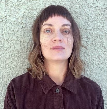 A woman with shoulder-length brown hair and bangs, wearing a dark brown shirt, standing against a textured wall.