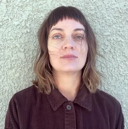 A woman with shoulder-length brown hair and bangs standing in front of a textured wall. She is wearing a dark, collared button-up shirt and has a neutral expression.