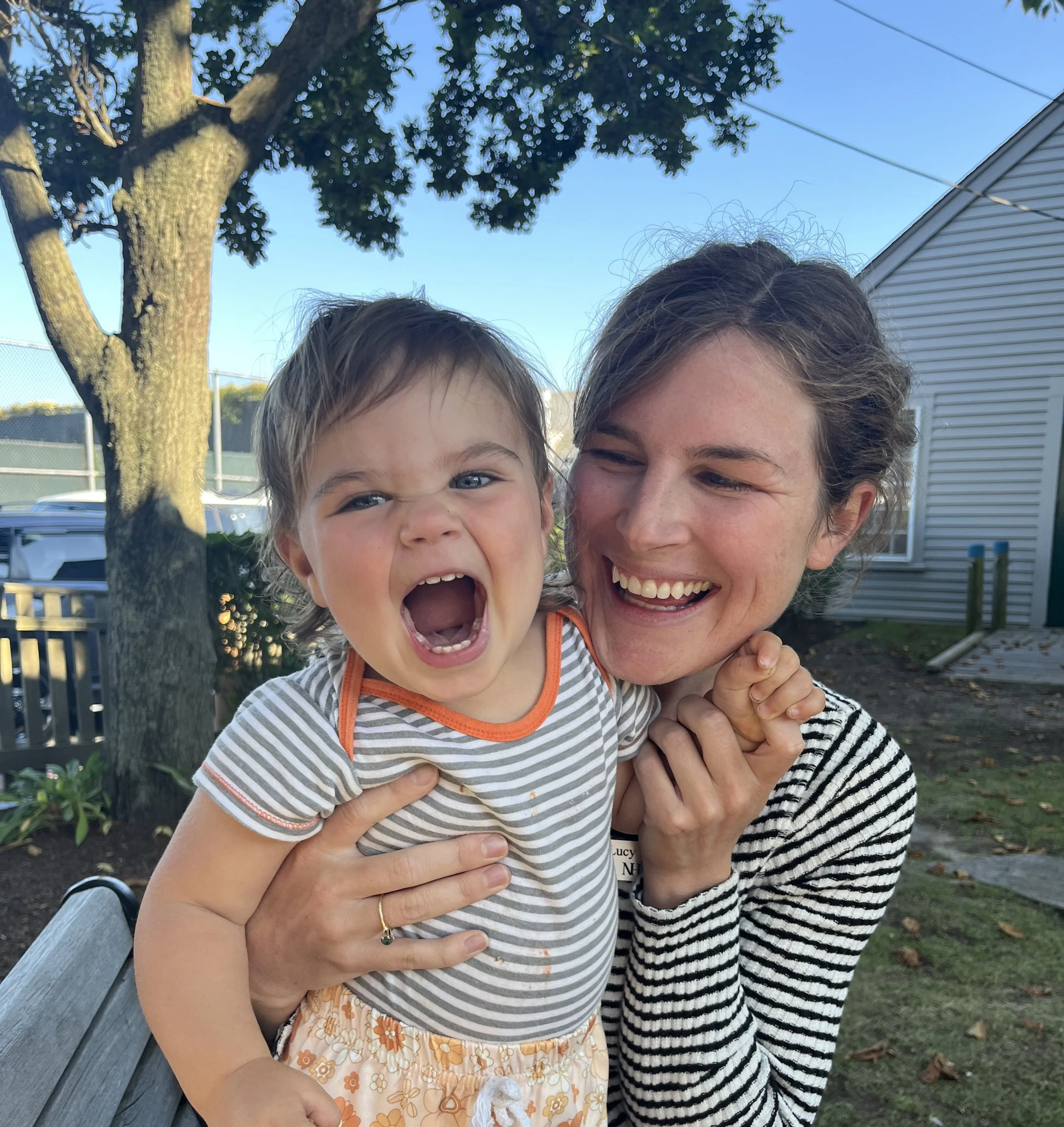 A woman and a young girl smiling outdoors, the woman hugging the girl, with trees, blue sky, and a house in the background.