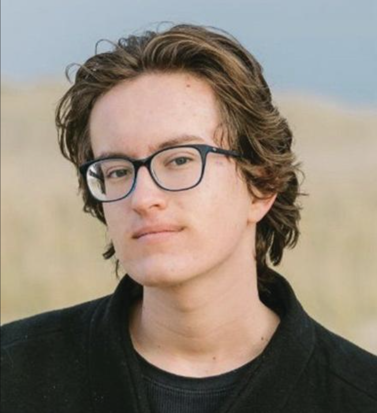 Portrait of a young person with brown, wavy hair, wearing glasses and a black shirt, outdoors with a blurry background of sand and sky.