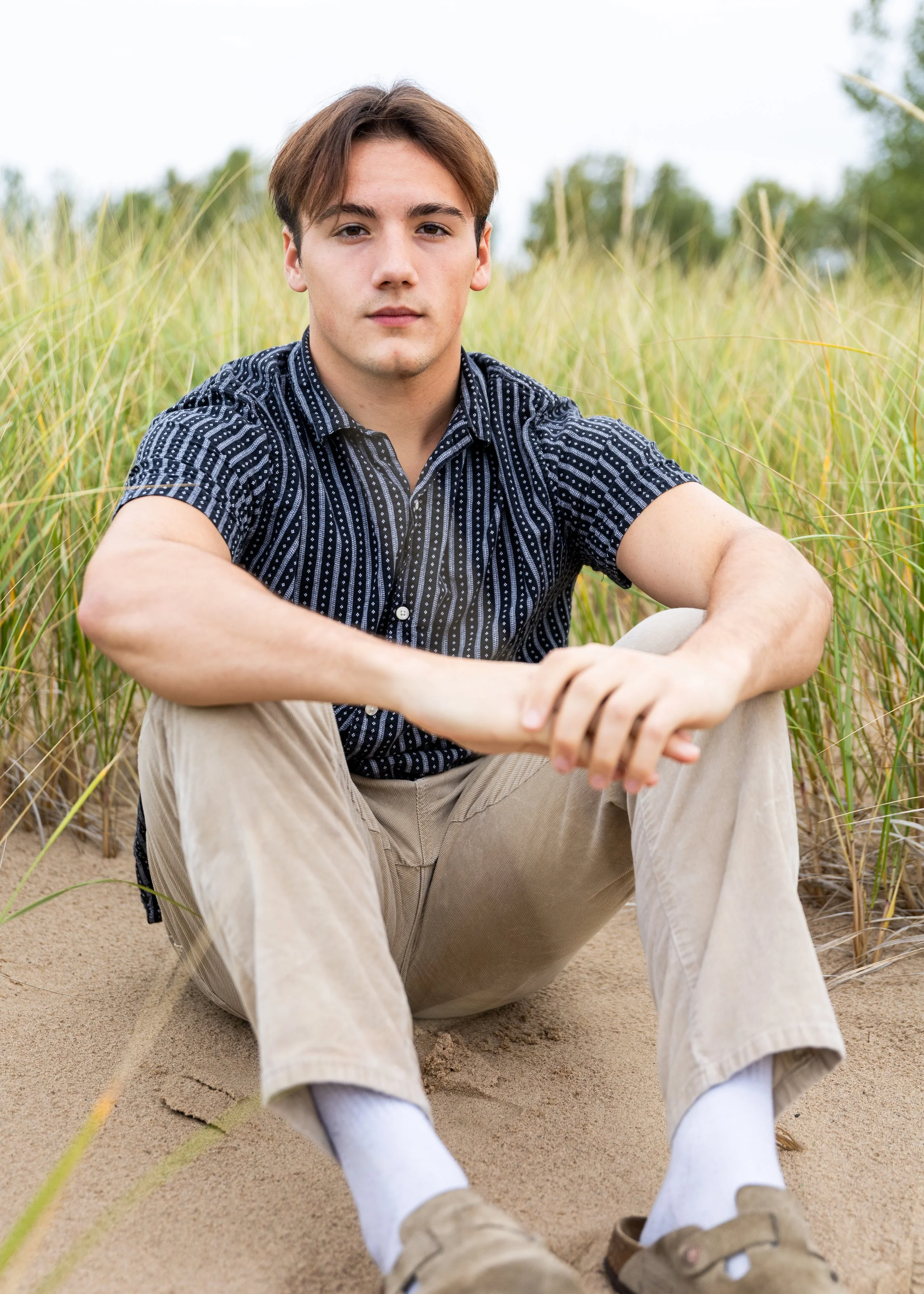 Young man sitting outdoors on sandy ground surrounded by tall grass, looking at camera with a neutral expression, wearing a patterned short-sleeve shirt, khaki pants, and white socks with beige shoes.