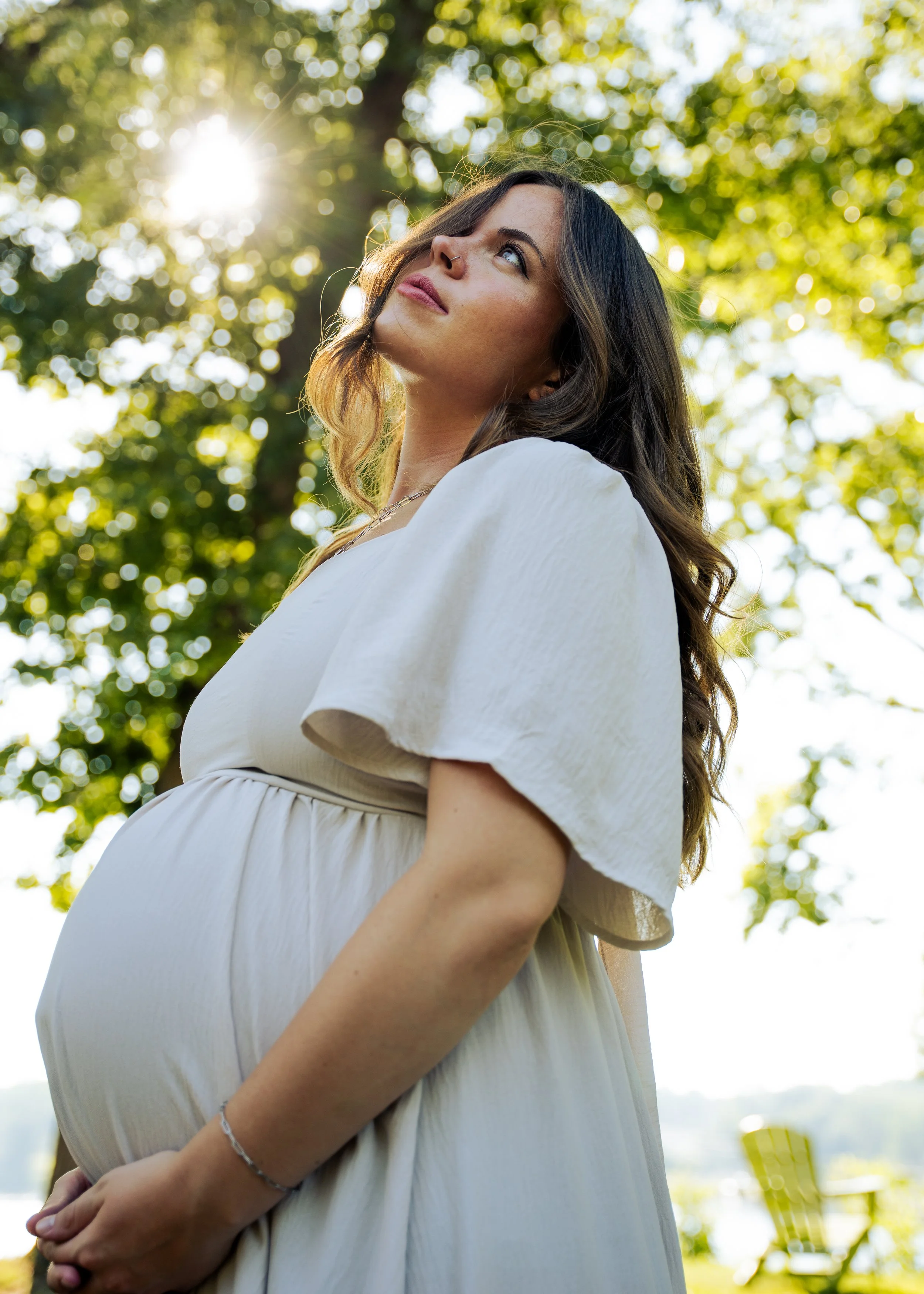 Pregnant woman standing outdoors in sunlight, gazing upward, with green trees and blurred outdoor furniture in the background.