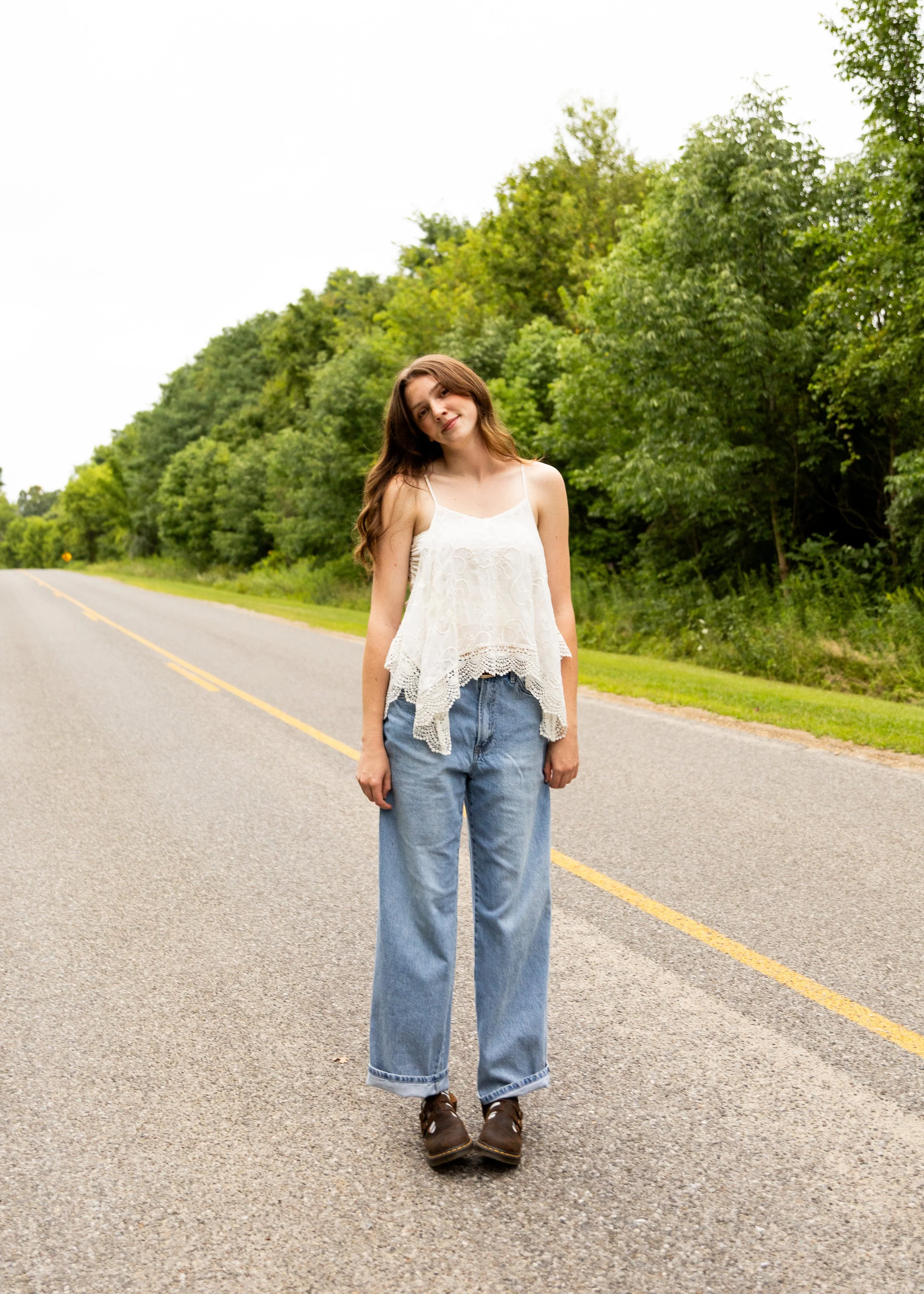 A young woman standing in the middle of a road with trees in the background, wearing a white sleeveless top and blue jeans.