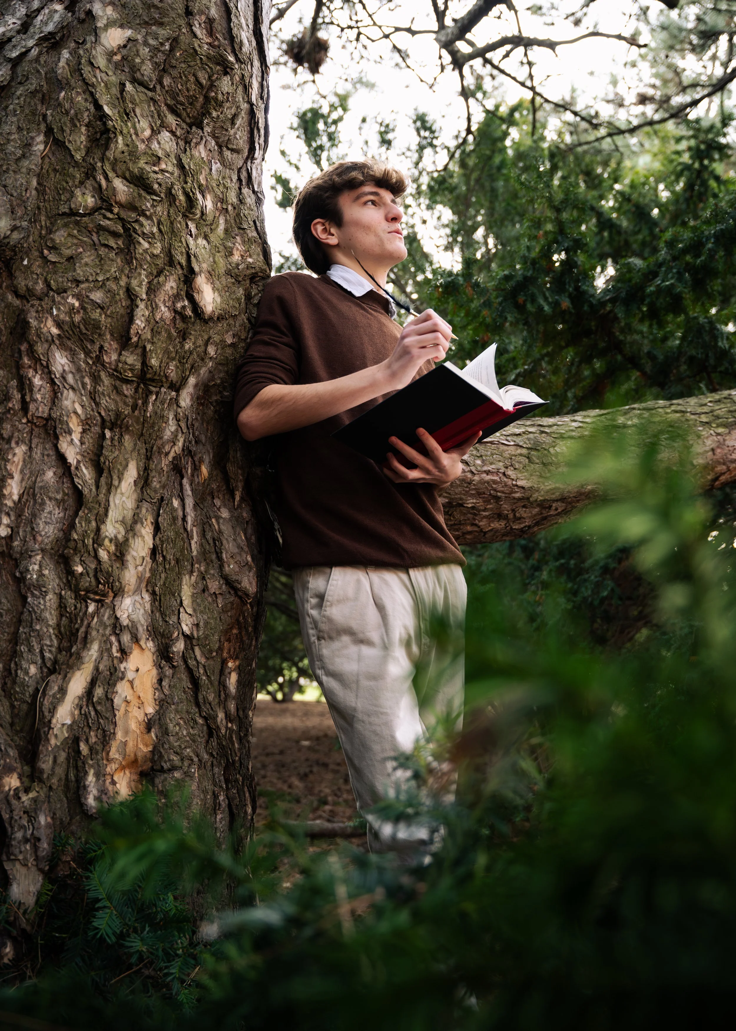 Young man leaning against a large tree with his eyes closed, holding an open book and a pen in his hand, surrounded by green foliage outdoors.