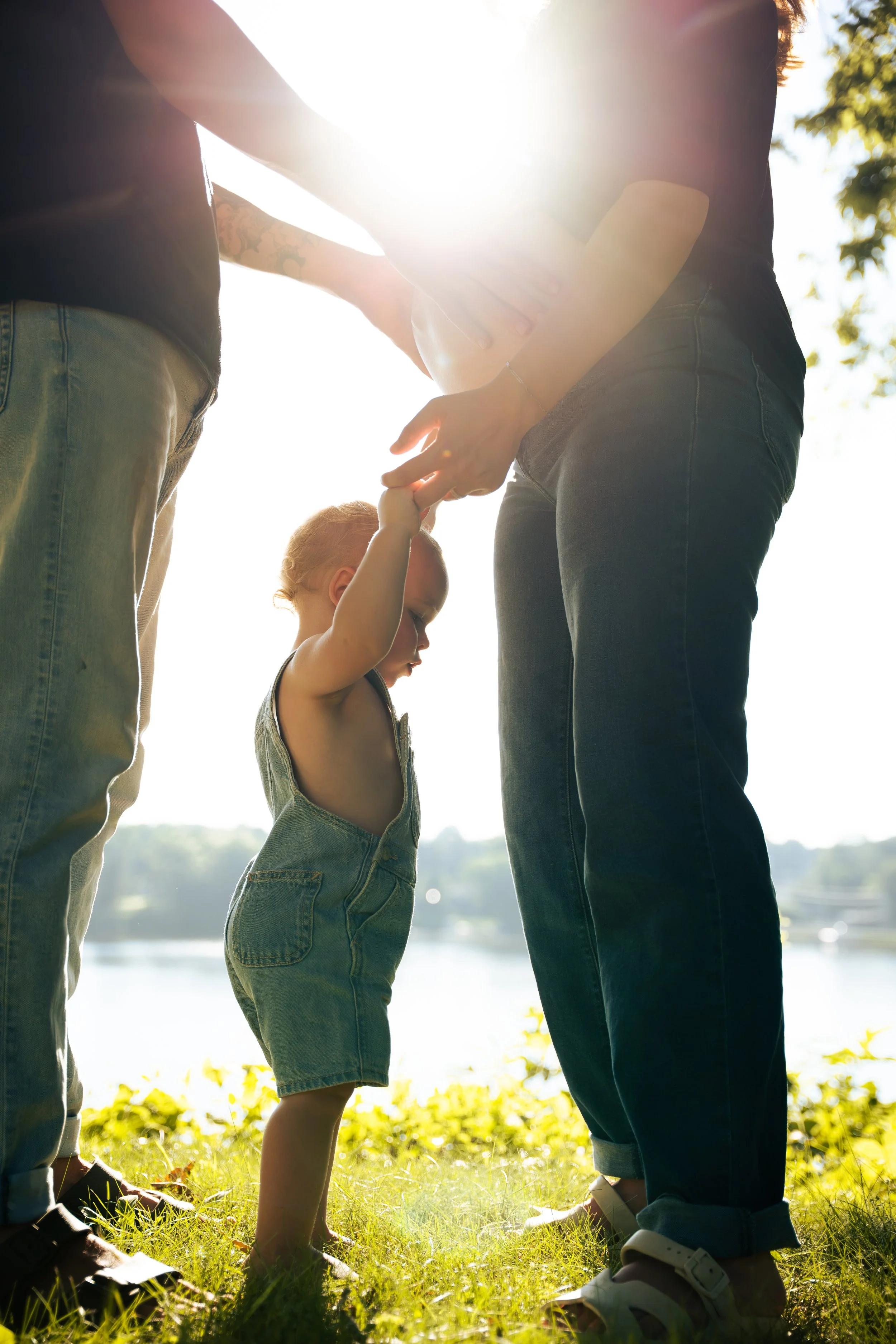 A young girl holding hands with two adults outdoors near a body of water, during sunny weather with sunlight shining through the trees.