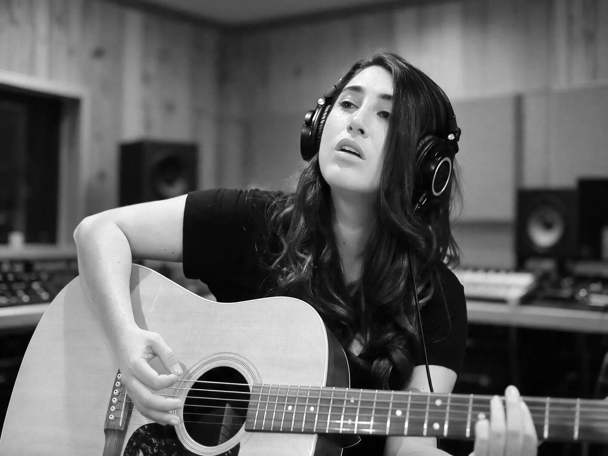 A woman with long hair is wearing headphones and playing an acoustic guitar in a music studio.