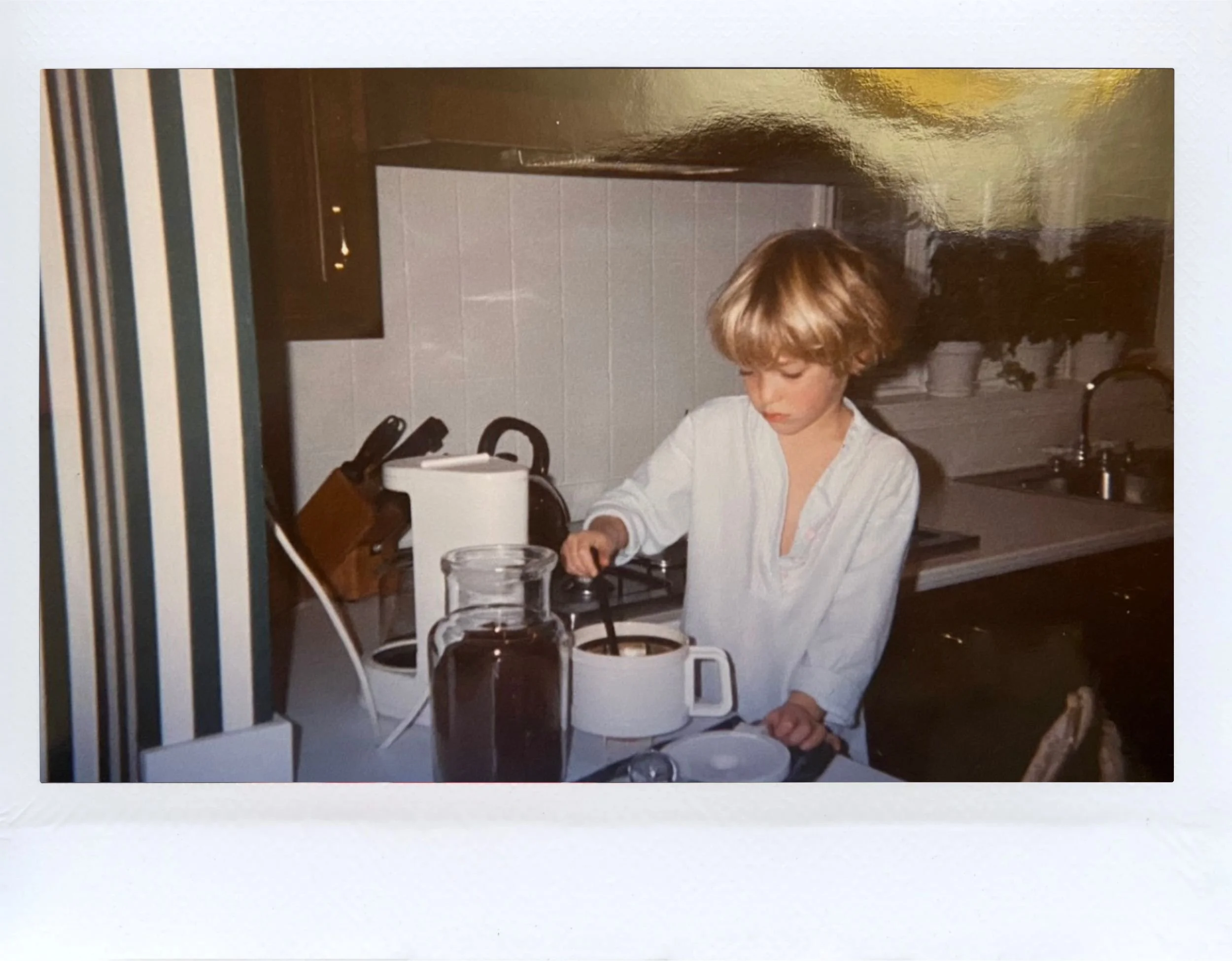 A young girl with blonde hair wearing a white shirt standing in a kitchen, preparing to cook on a stovetop with a coffee maker and glass pitcher on the counter.