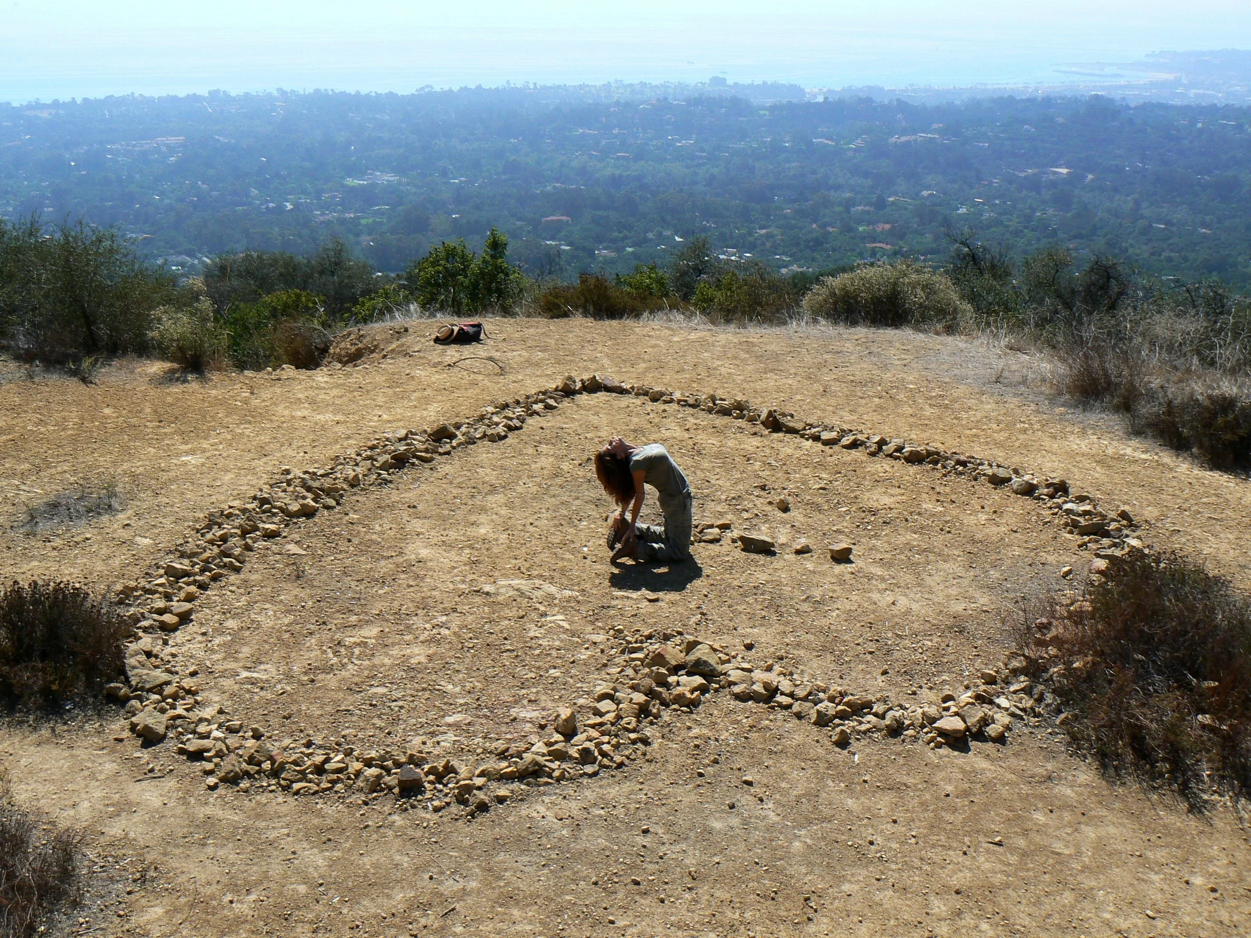 Lisa is in a yoga posture outside in the center of a large heart shape formed with stones with a panoramic view.