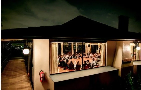 Indoor event with people seated, viewed from outside through large windows at night.