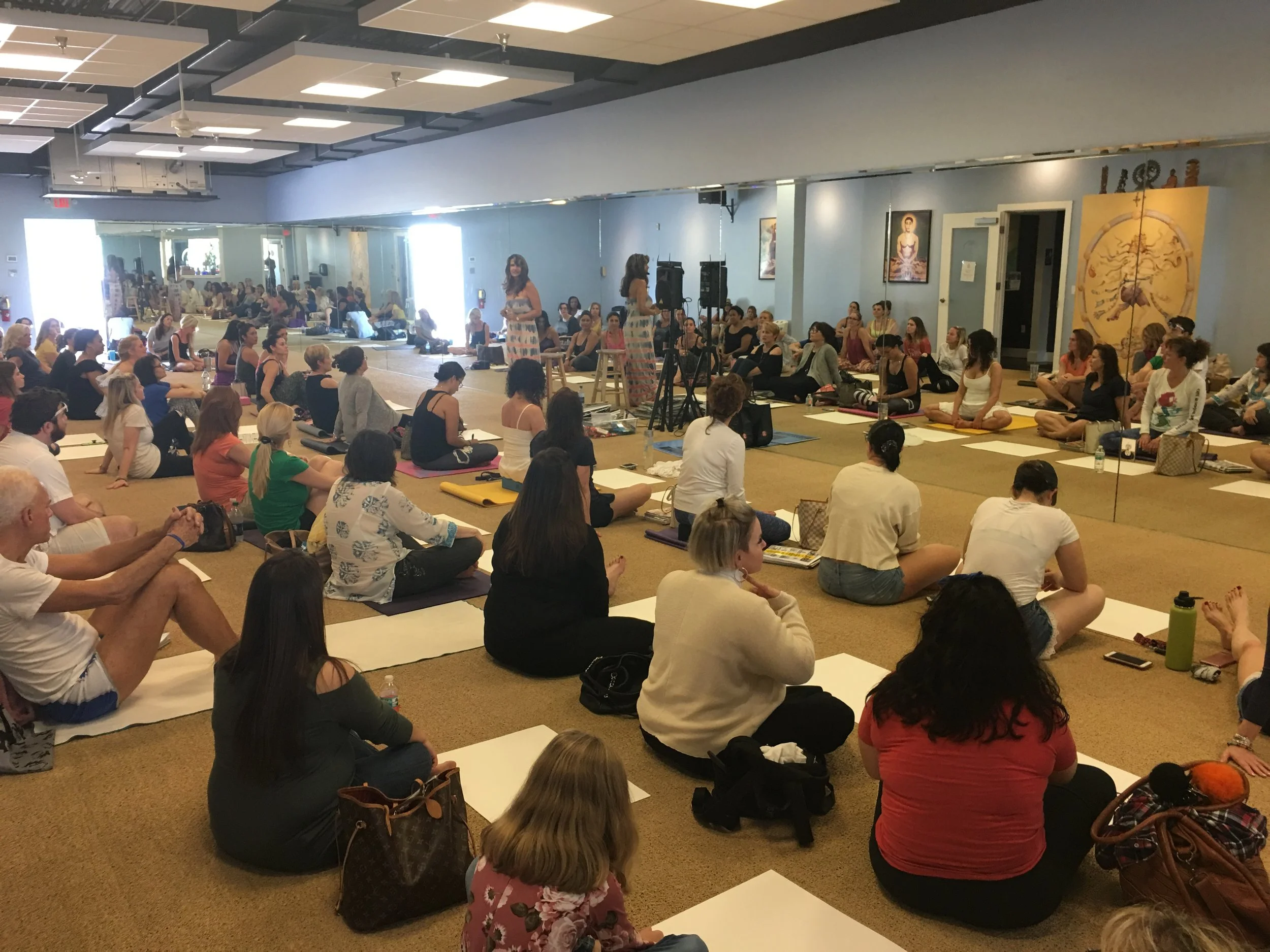 A large group of people seated on the floor in a spacious room attending a workshop or seminar. Lisa is standing at the front, leading the session, with music equipment and artwork on the walls.