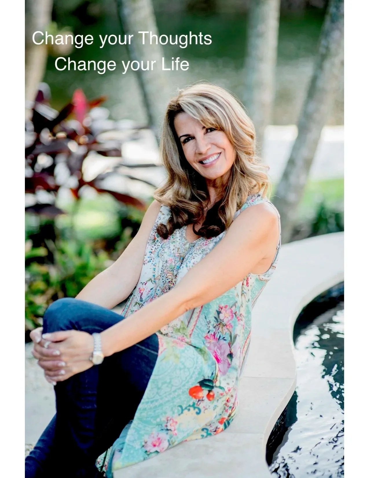 Lisa Goodwin pictured with long, wavy blonde hair smiling, sitting beside a small pond or water feature in a park with trees in the background, wearing a sleeveless floral top and dark pants.