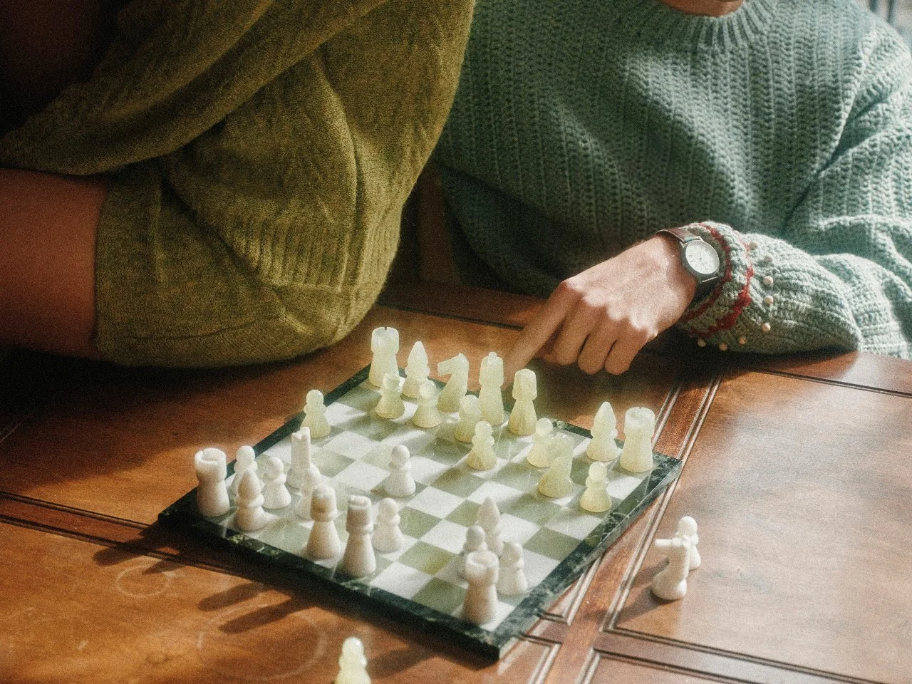 Two people playing chess on a wooden table, with a glass chess set, one person pointing at a piece, both wearing cozy sweaters.