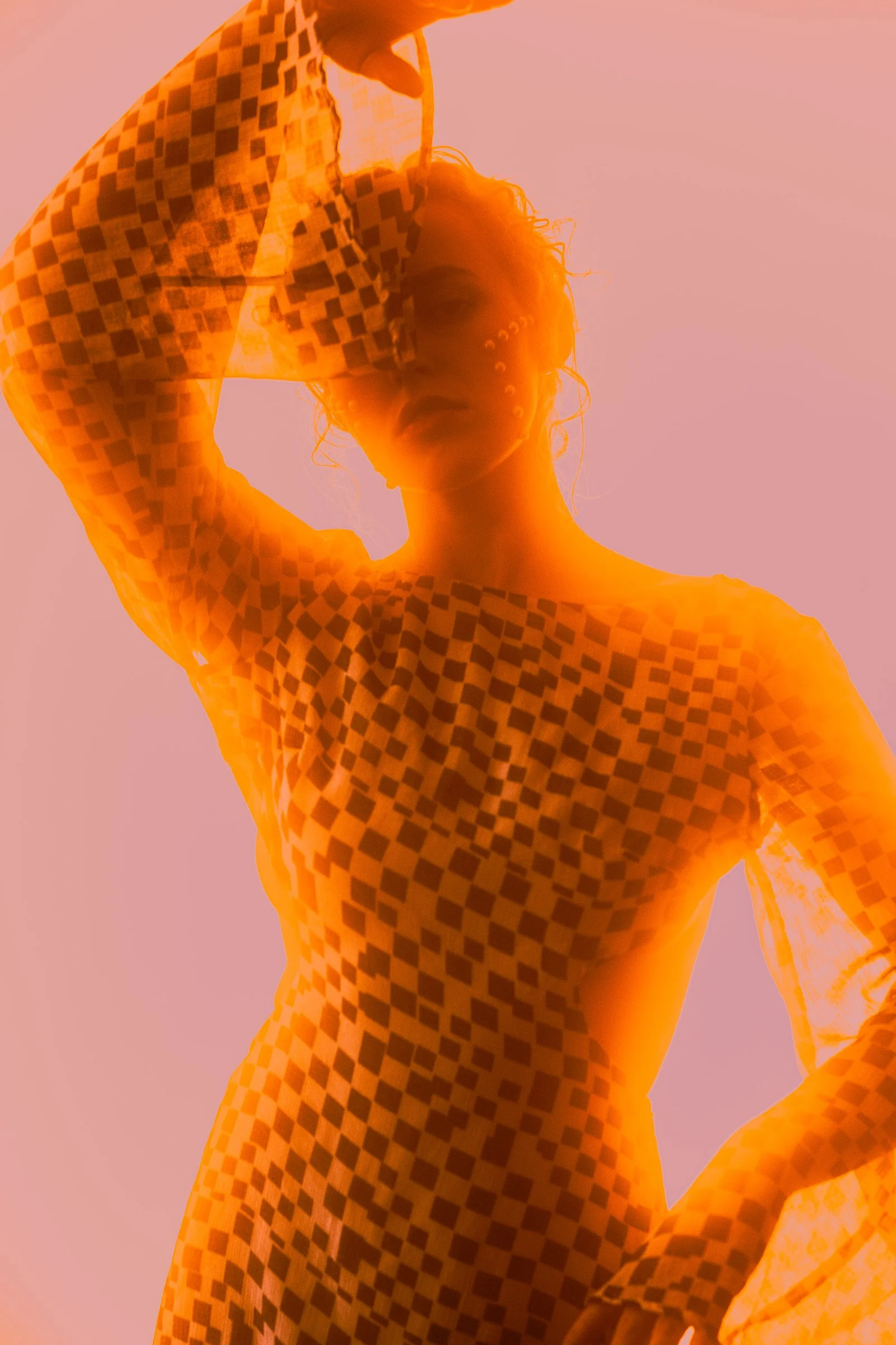 A woman with curly hair and face partially covered with a face ring, posing with her hand behind her head, wearing a checkered long-sleeve top, against a pink background illuminated with orange light.