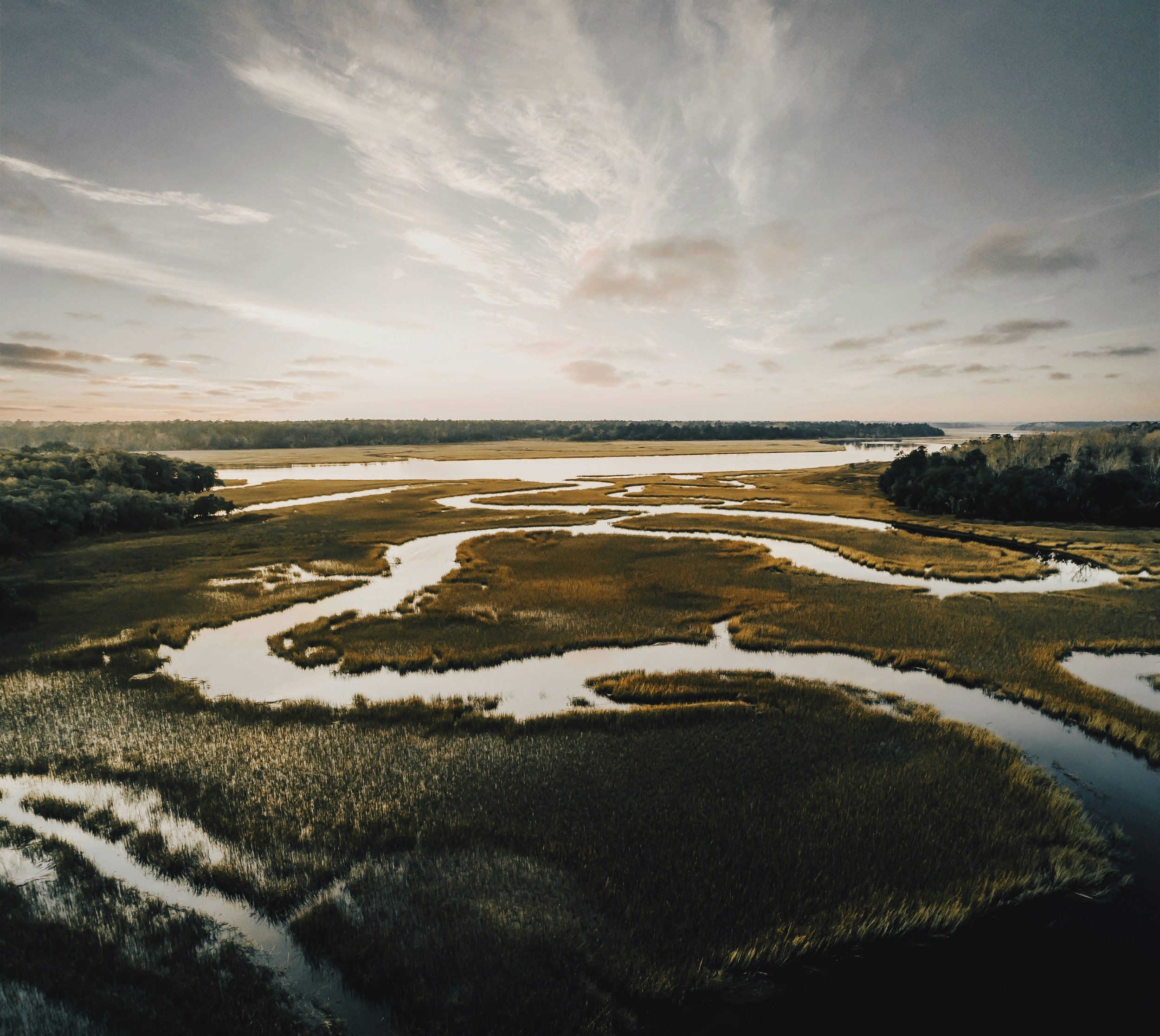A landscape view of a winding river through a grassy area under a partly cloudy sky during sunset.