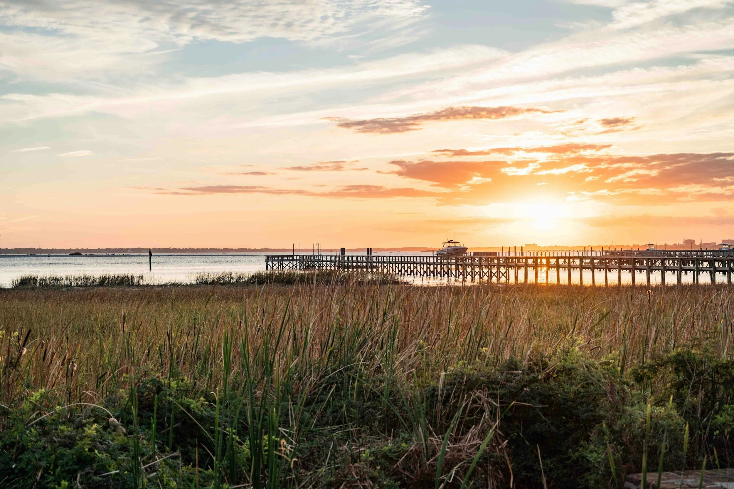 Sunset over a water body with a wooden pier, a boat, grassy foreground, and a sky with scattered clouds.