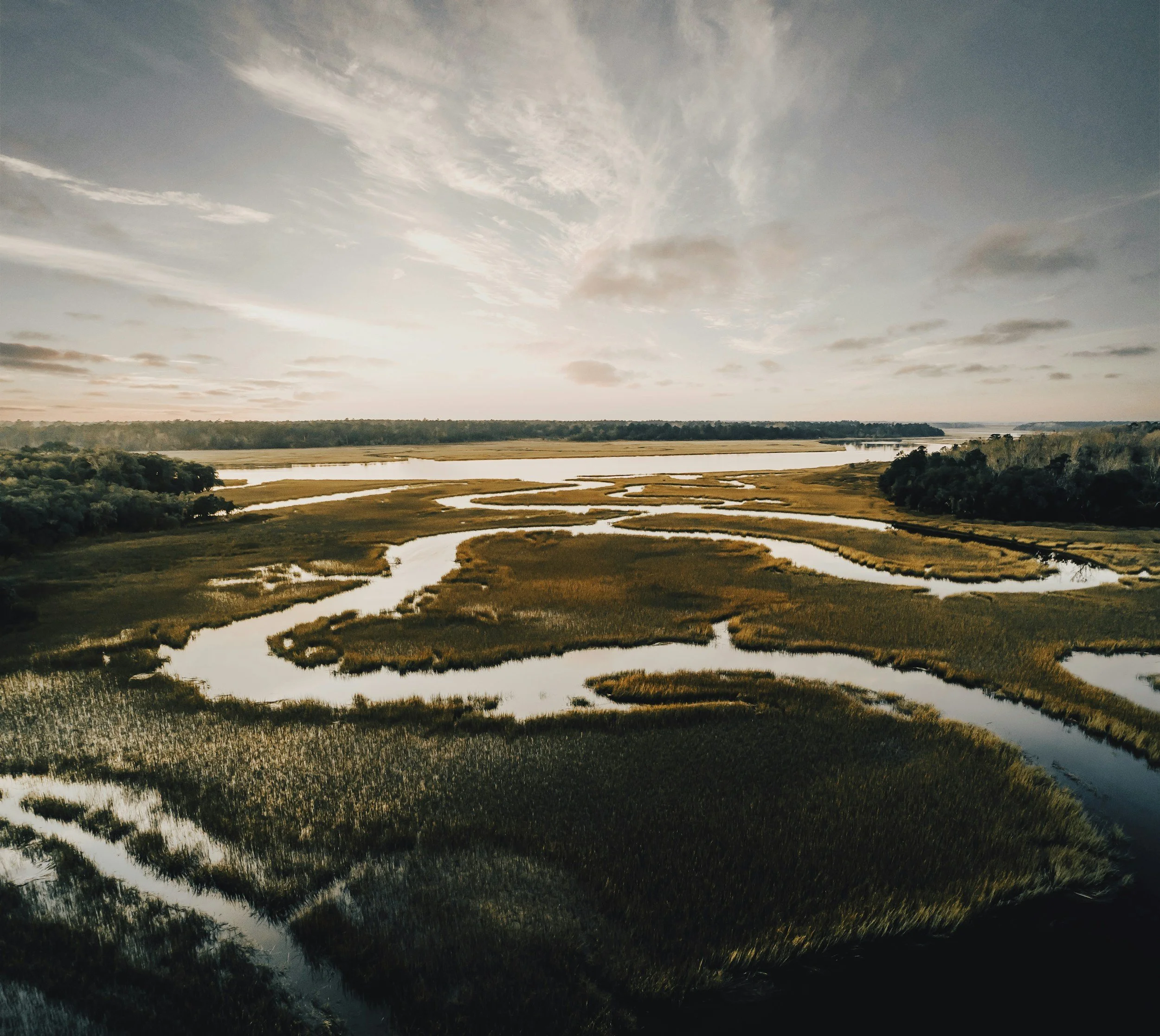 A wide view of a river winding through a landscape with marshes and trees, under a partly cloudy sky during sunset or sunrise.