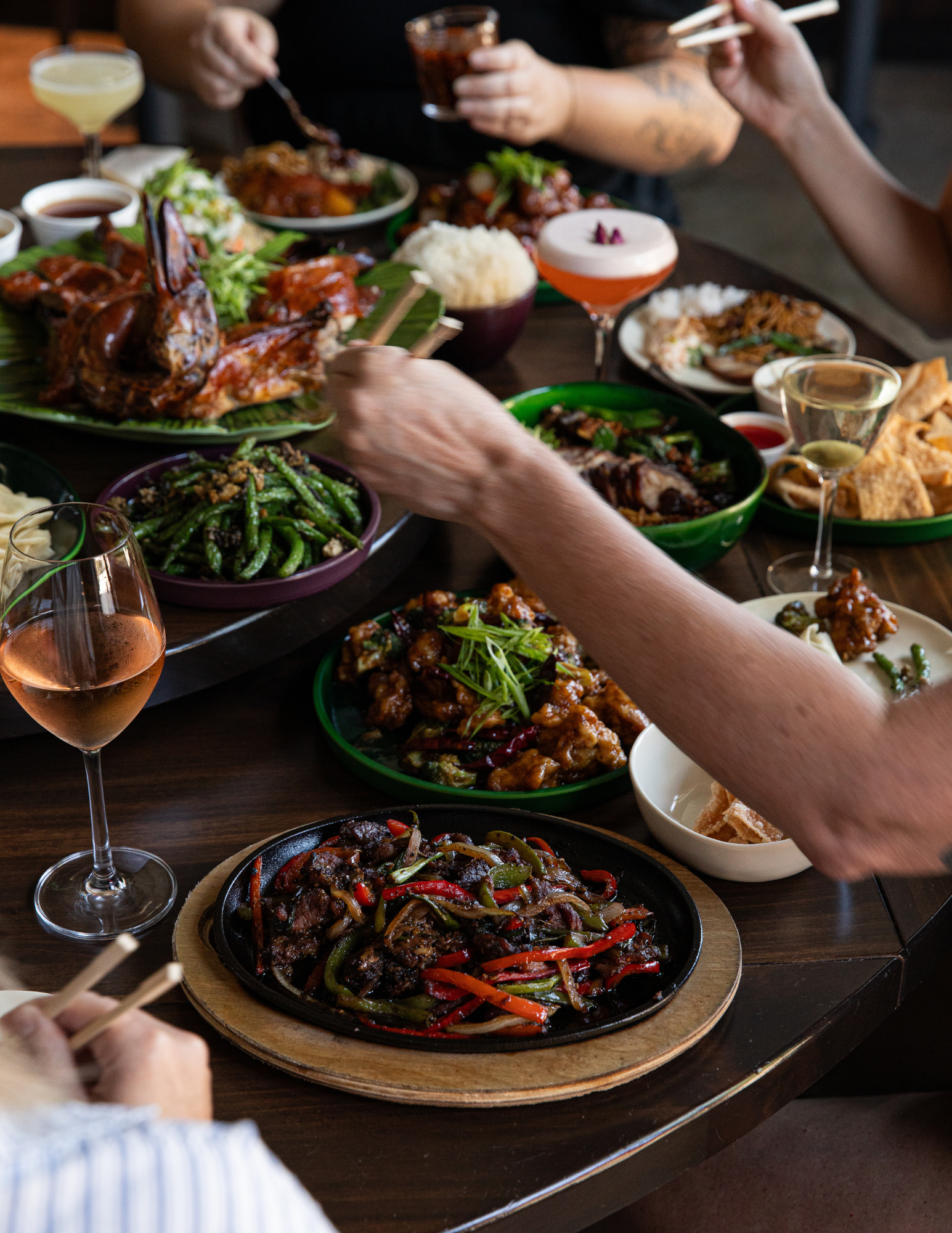 A table filled with various Chinese dishes, including stir-fried vegetables and meats, rice, and sauces, with people using chopsticks and holding drinks at Beautiful South by Kwei Fei in downtown Charleston, South Carolina