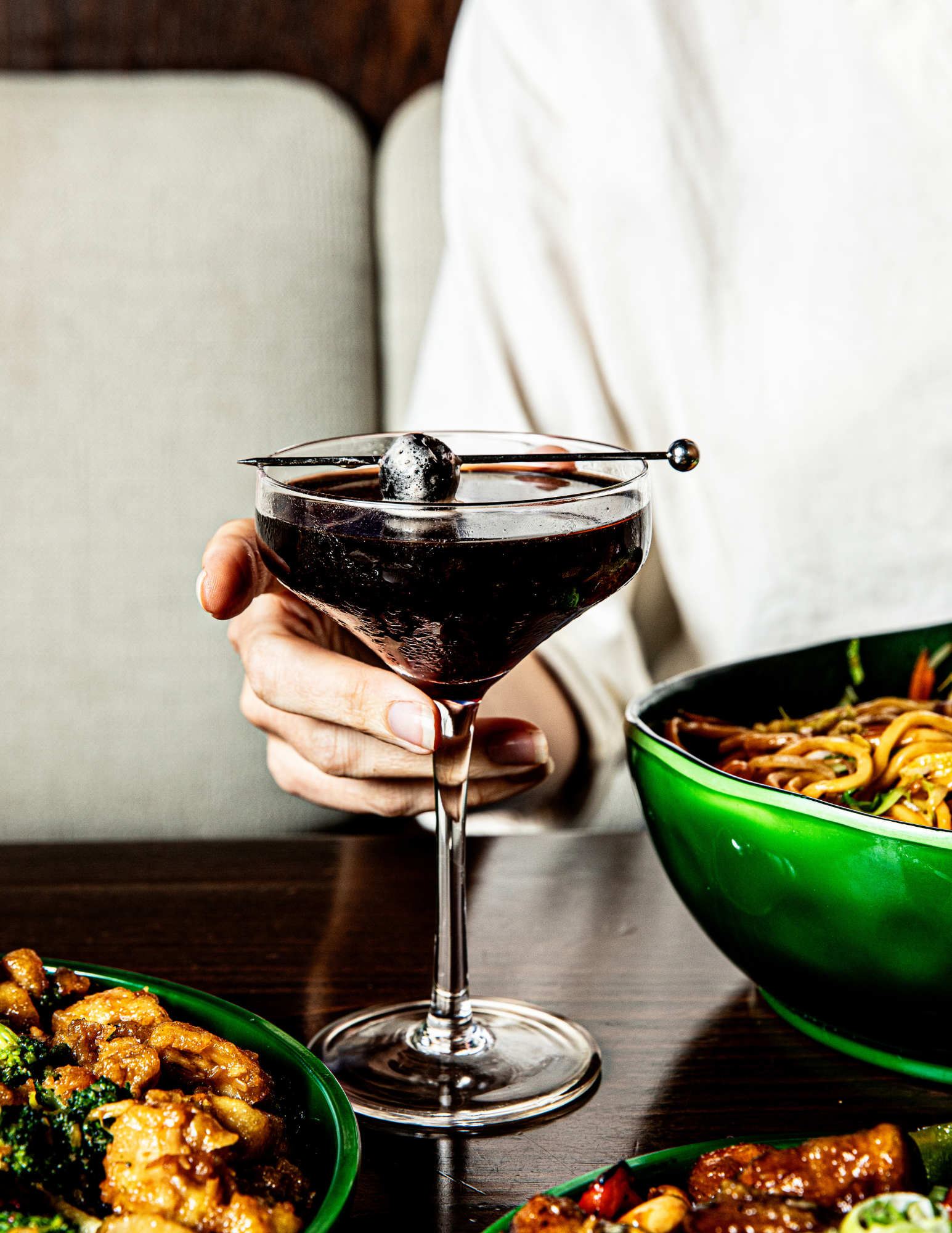 A person holding a espresso martini with a black cherry garnishing, surrounded by bowls of Asian-style stir-fry dishes on a dark wooden table at Beautiful South by Kwei Fei in downtown Charleston, South Carolina
