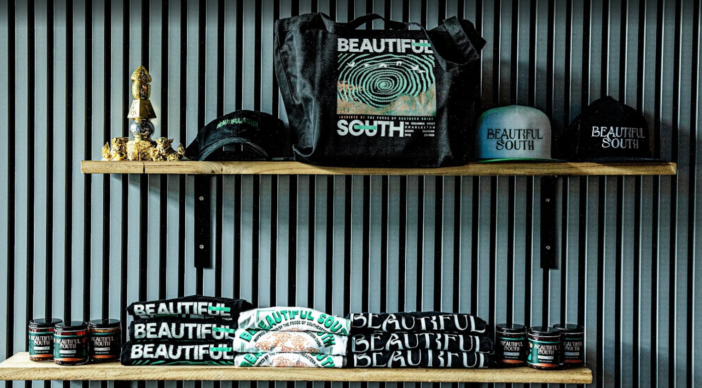 Display of merchandise on two wooden shelves against a corrugated metal wall, including hats, a tote bag, T-shirts, and small containers, all branded with "Beautiful South."