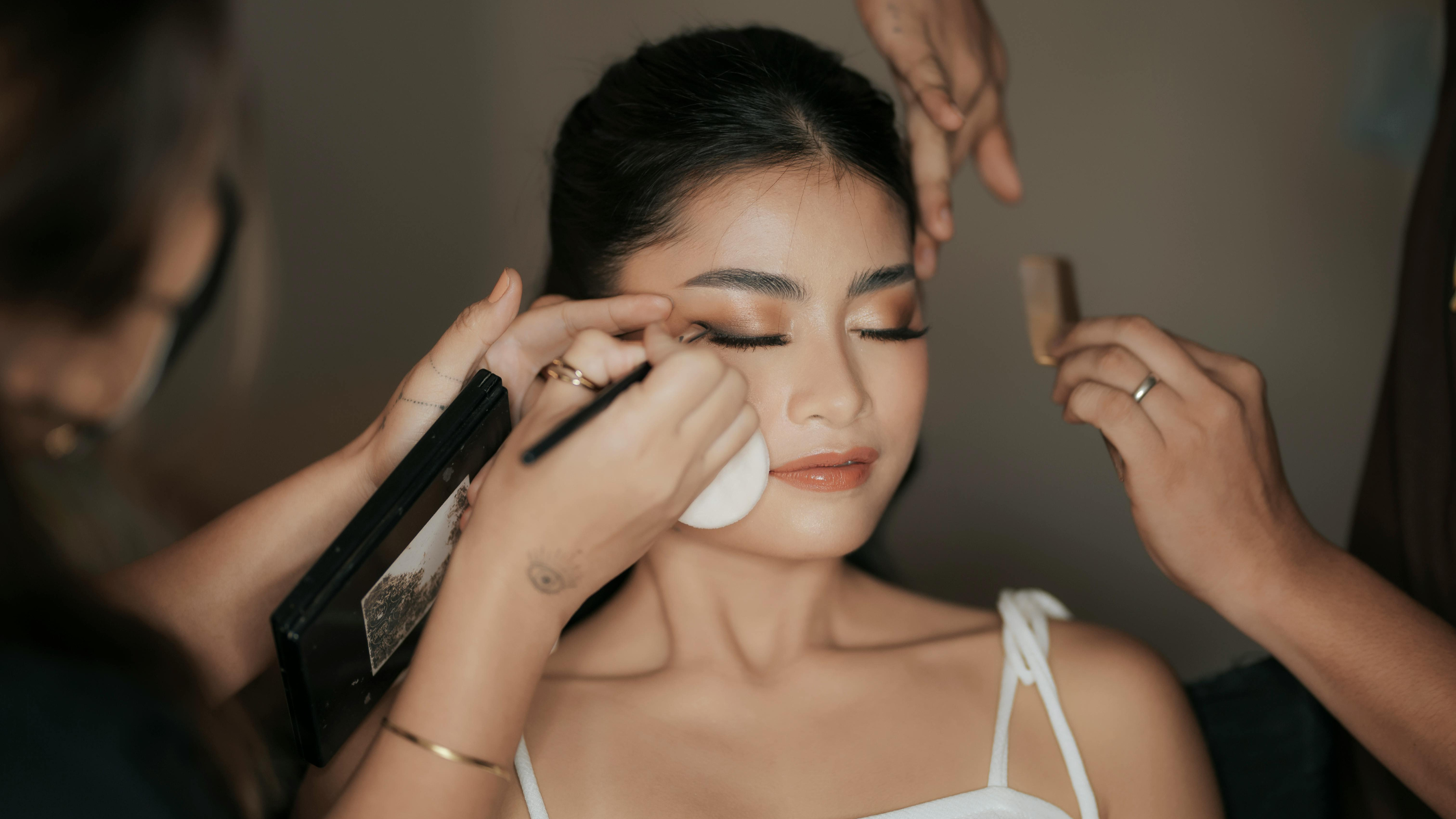 Makeup artist applying eyeliner to a woman with closed eyes, dark hair, and a white top, in a beauty studio.