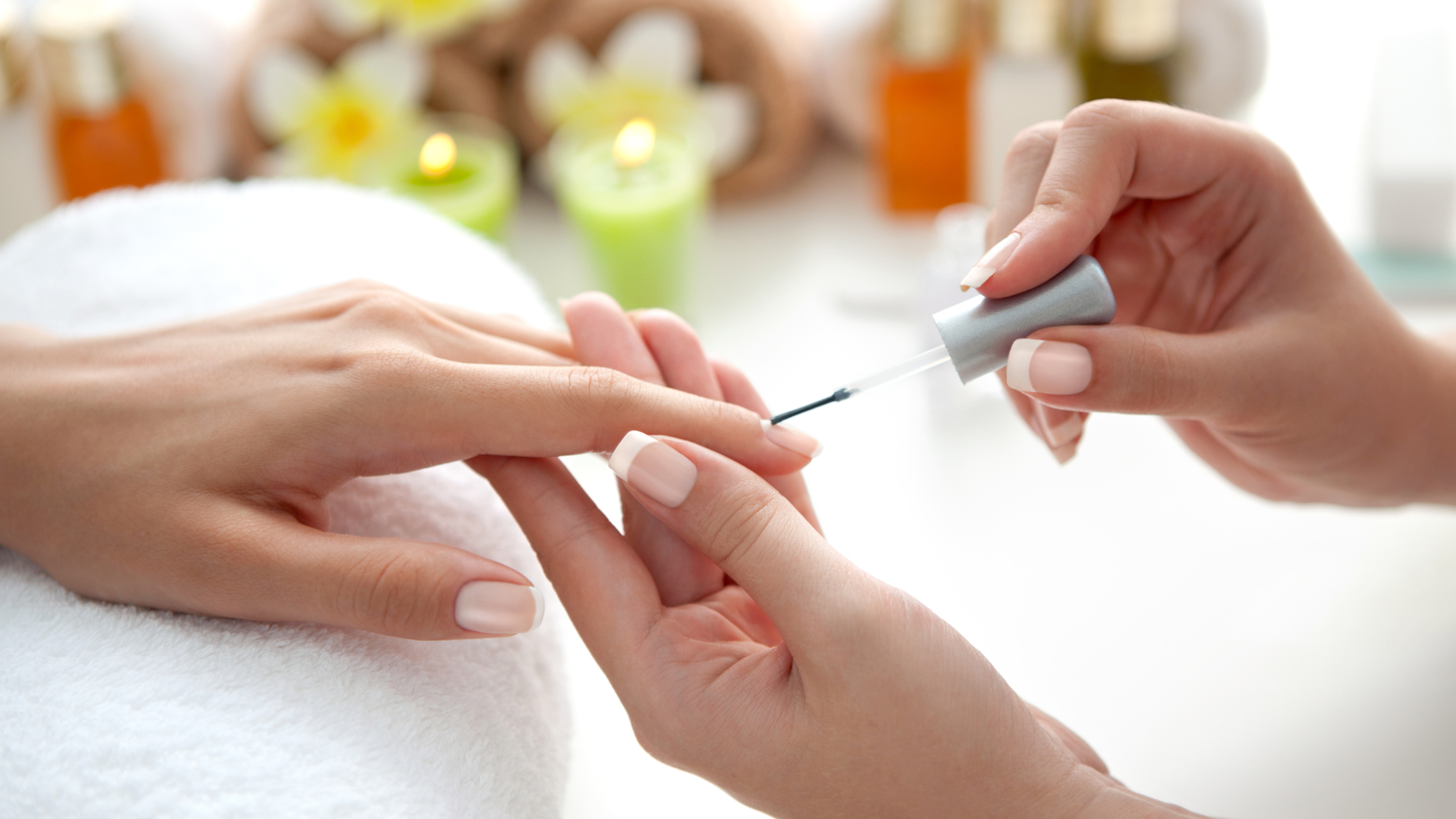 A person receiving a manicure with clear nail polish, with candles and bottles in the background.
