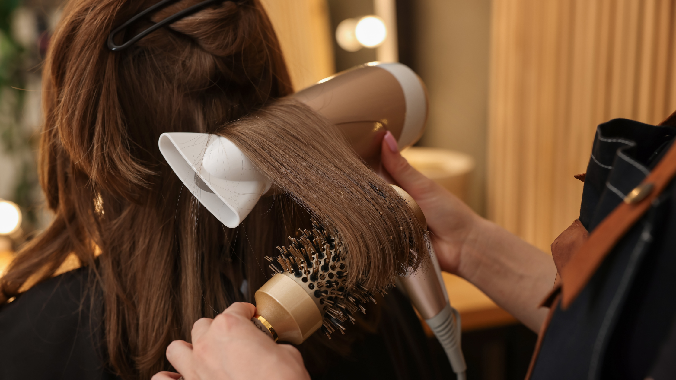 Person blow-drying long brown hair with a round brush and hairdryer in a salon.